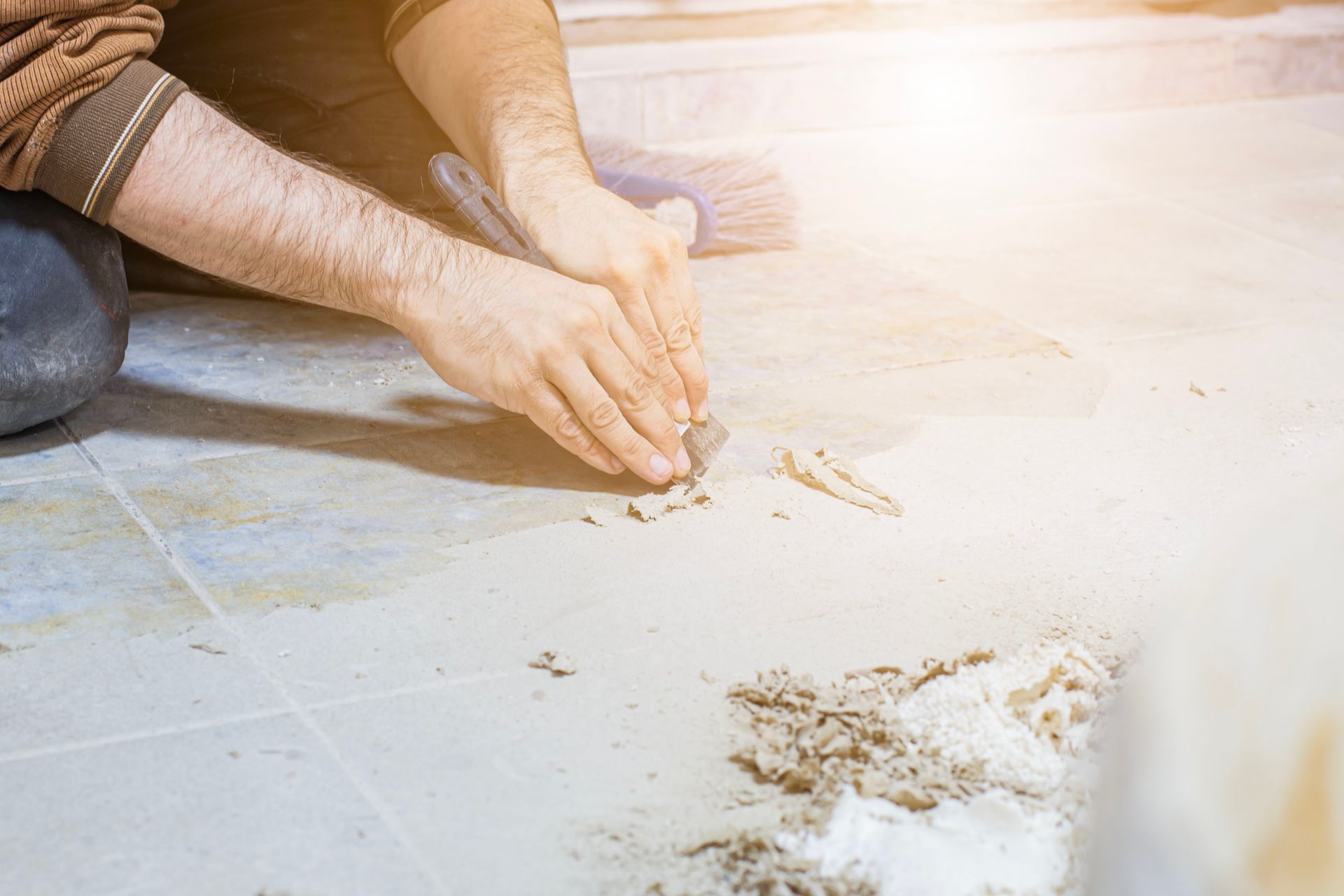 Hands using a utility knife to scrape residue from a tiled floor during a renovation.