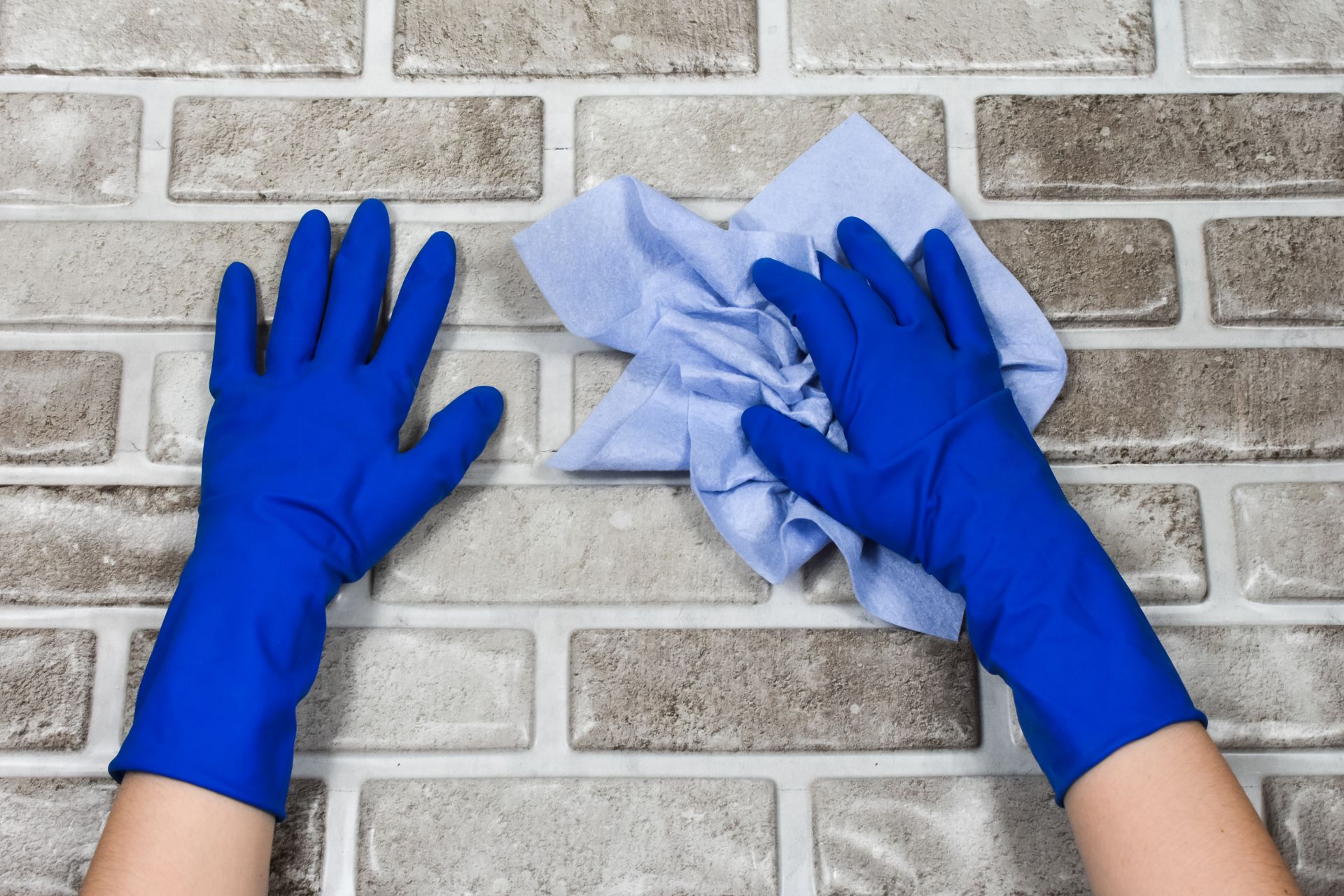 Hands wearing bright blue rubber gloves are cleaning a light-colored, faux-brick textured wall with a light blue cloth.
