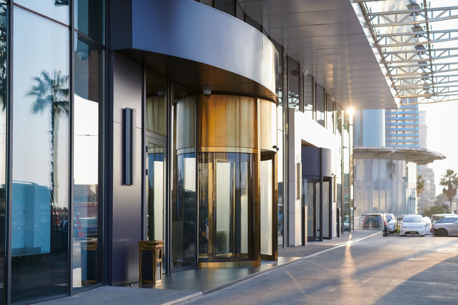 A modern glass facade with a gold-trimmed revolving door at a building entrance, with parked cars on a sunny street.