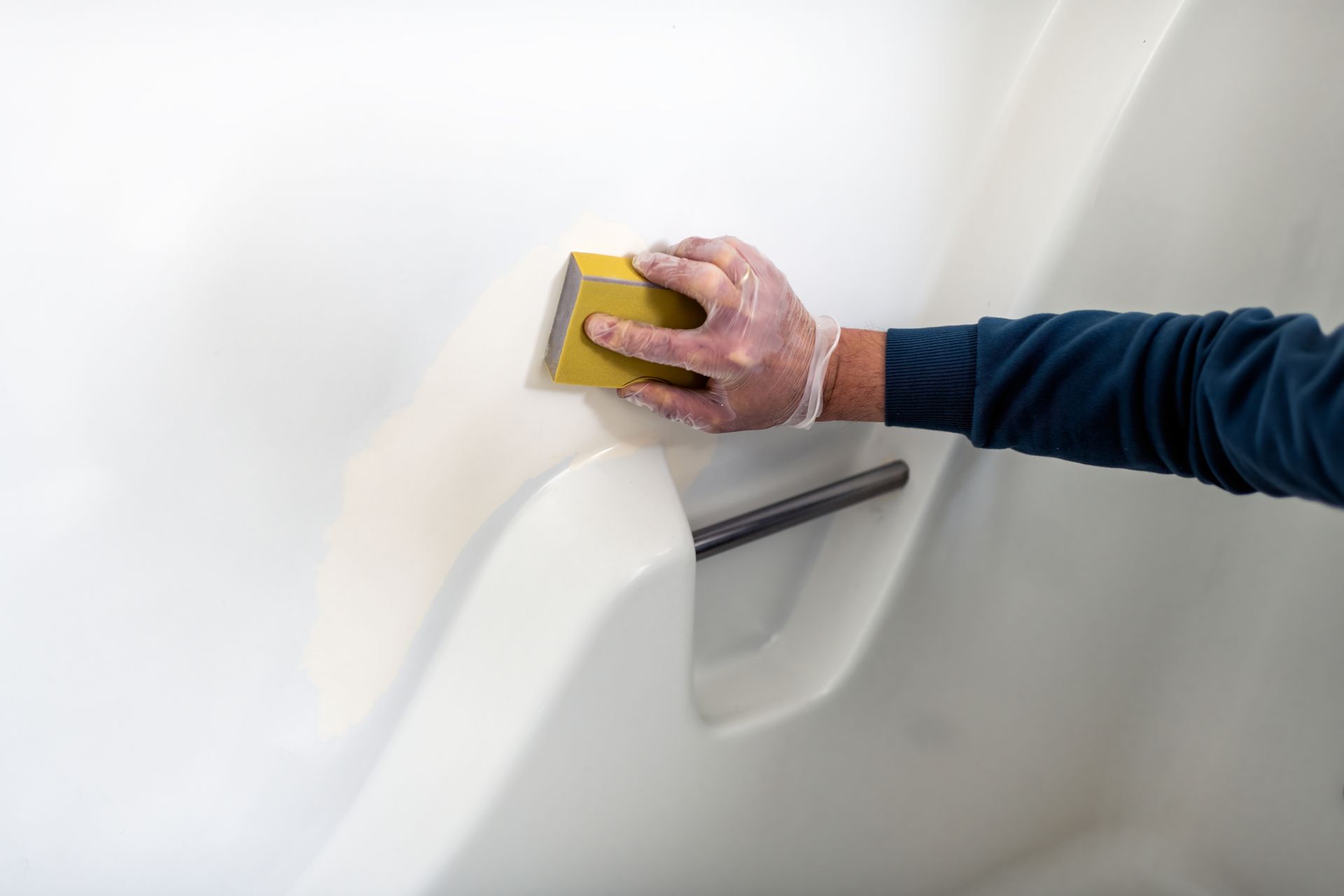 A gloved hand uses a yellow sanding block to smooth the surface of a white bathtub enclosure.
