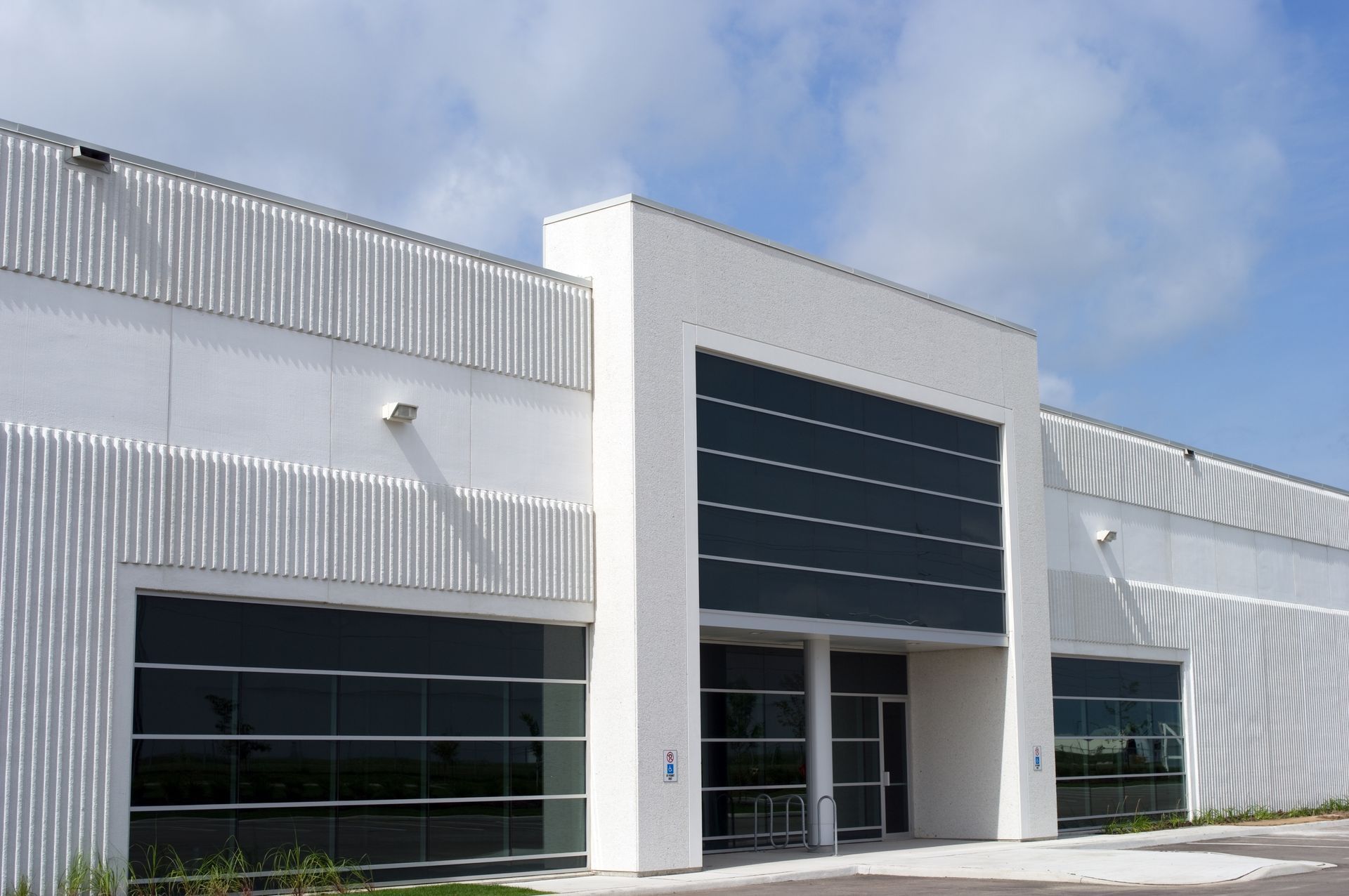 Modern white commercial building with glass paneled doors and windows under a partly cloudy blue sky.