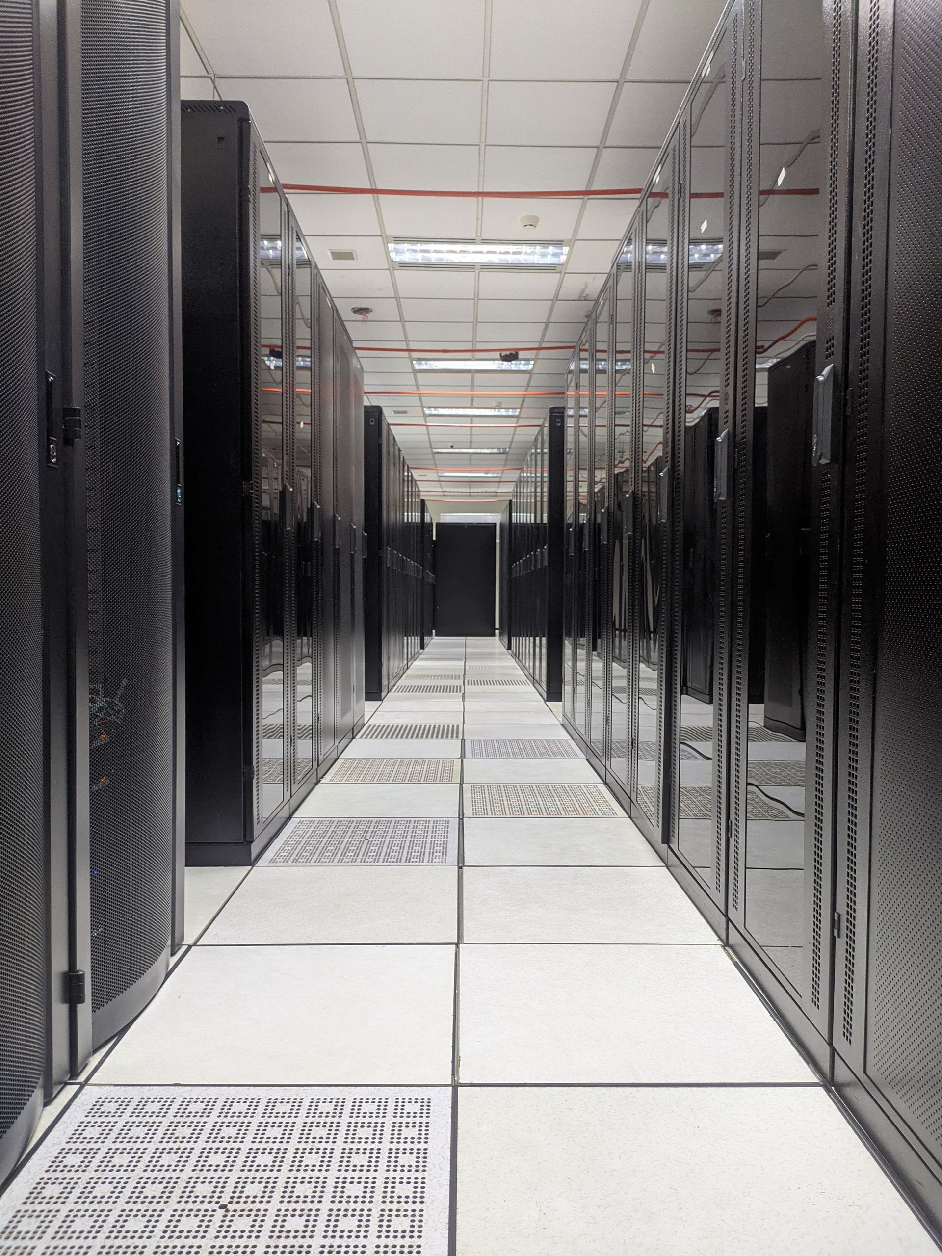 A long aisle in a data center with rows of black server racks and white tiled flooring