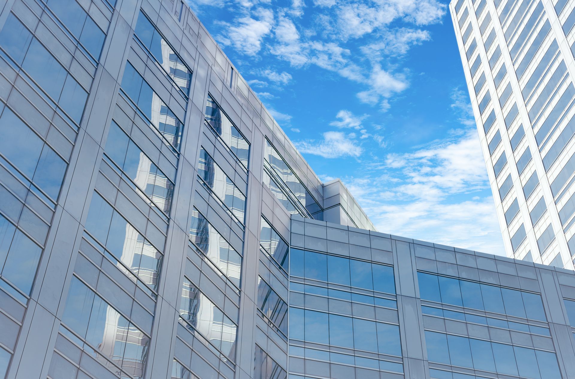 A low-angle view of modern, glass-paneled office buildings under a bright blue, partly cloudy sky.