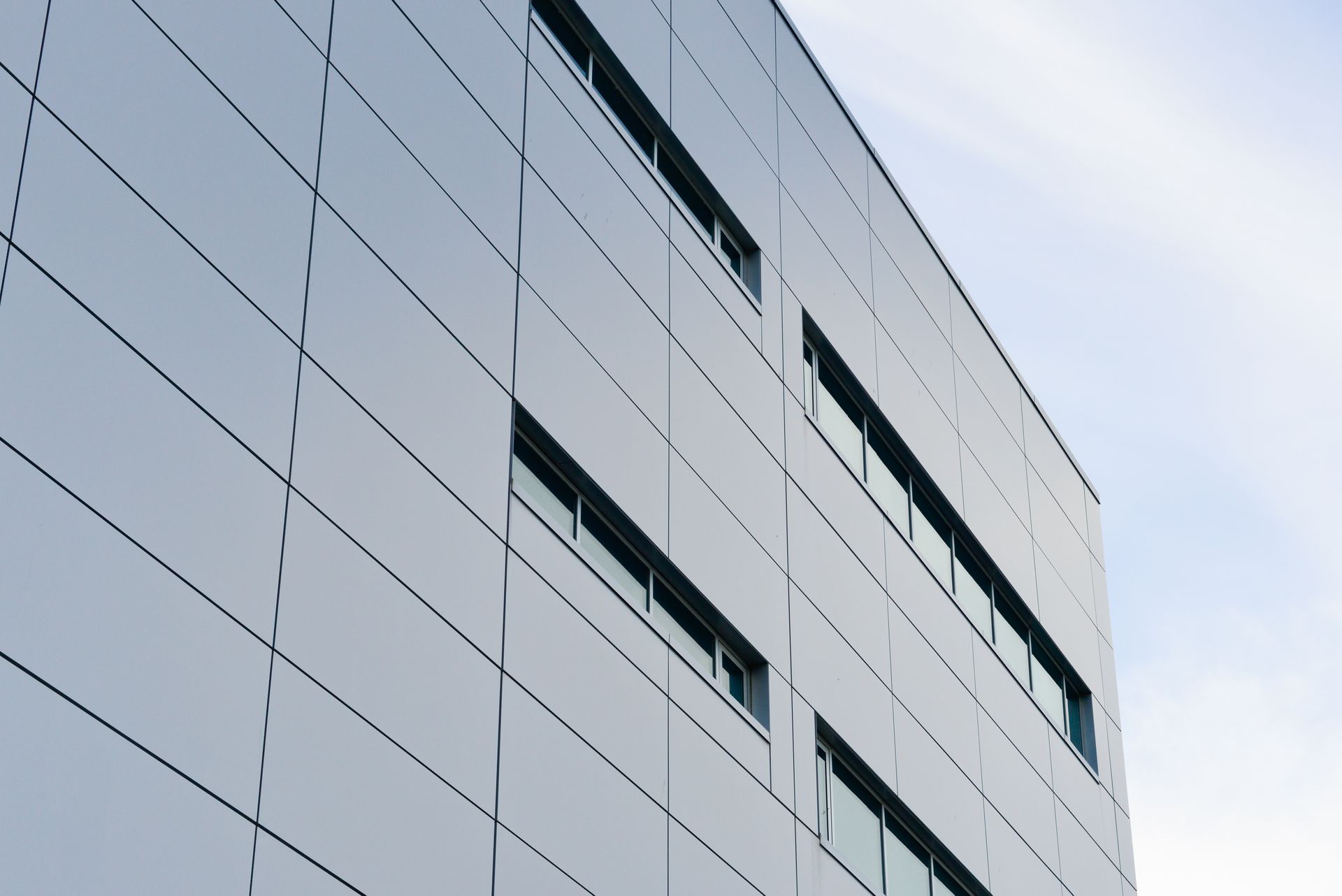 A low-angle view of a modern building facade covered in gray, rectangular metal panels with several horizontal windows.