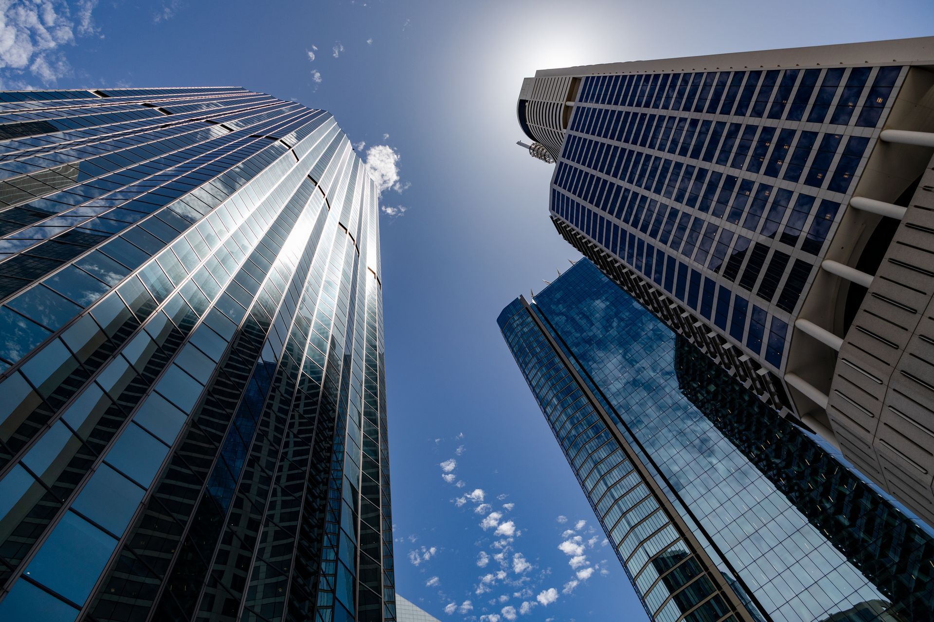 Low-angle view of three modern glass skyscrapers against a bright blue, sunny sky with light clouds.