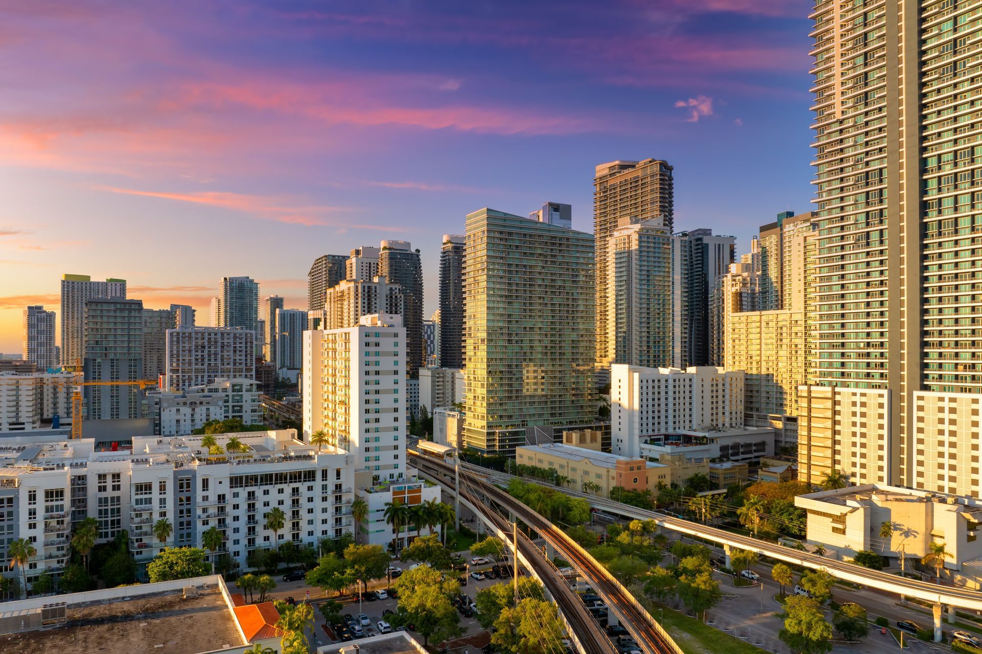 A sunset casts a golden glow over a modern city skyline with high-rise buildings and an elevated train track.