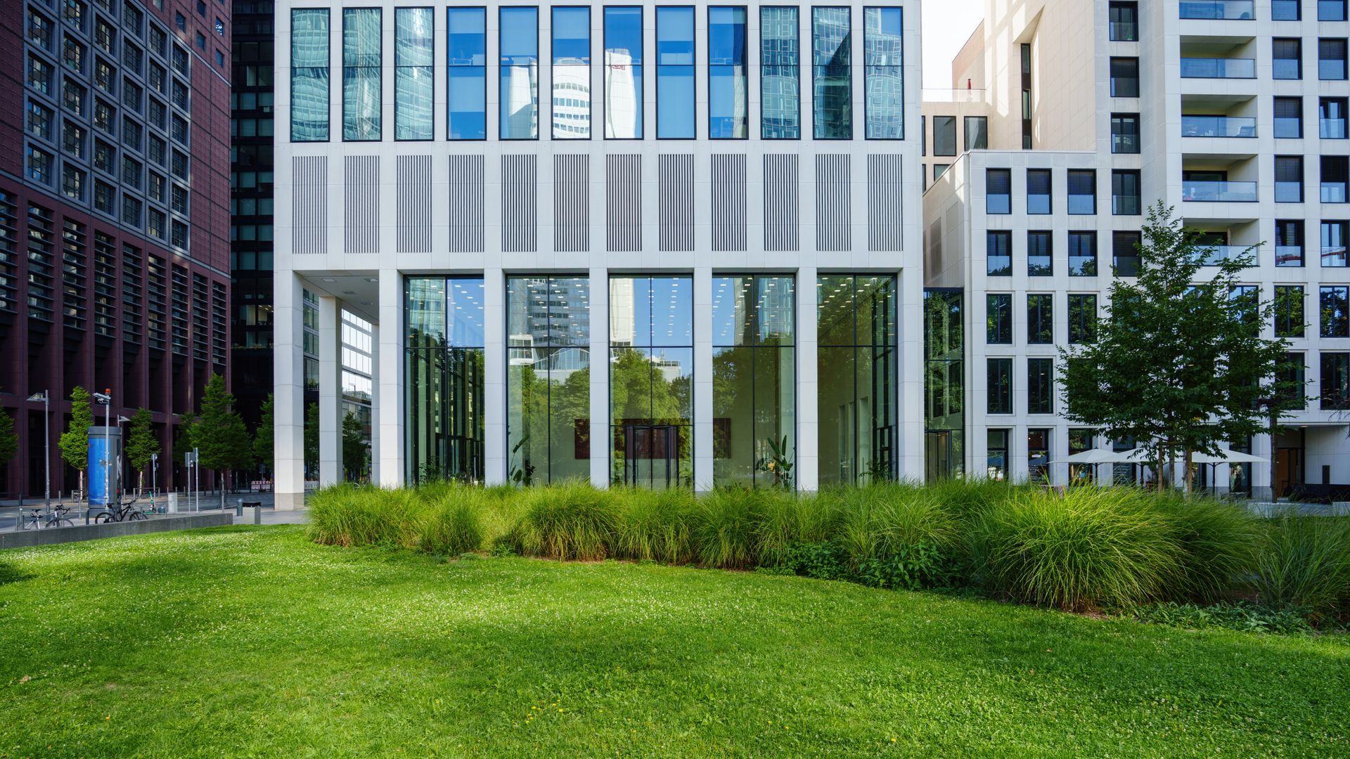 A modern white building with large windows stands behind a lush green lawn and bushes in a city setting.