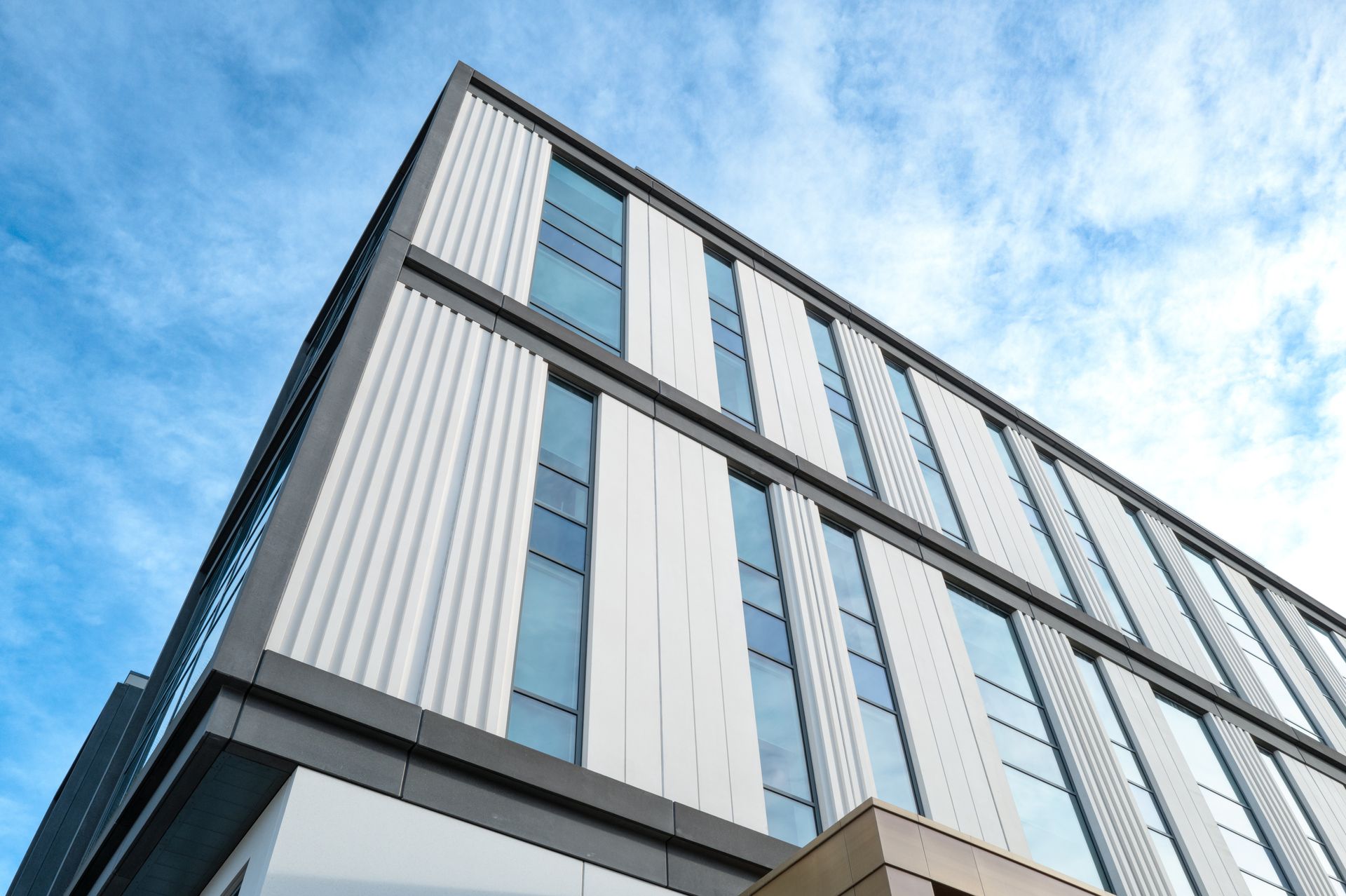 Low-angle view of a modern building with white vertical panels and dark grey frames against a blue, cloudy sky.
