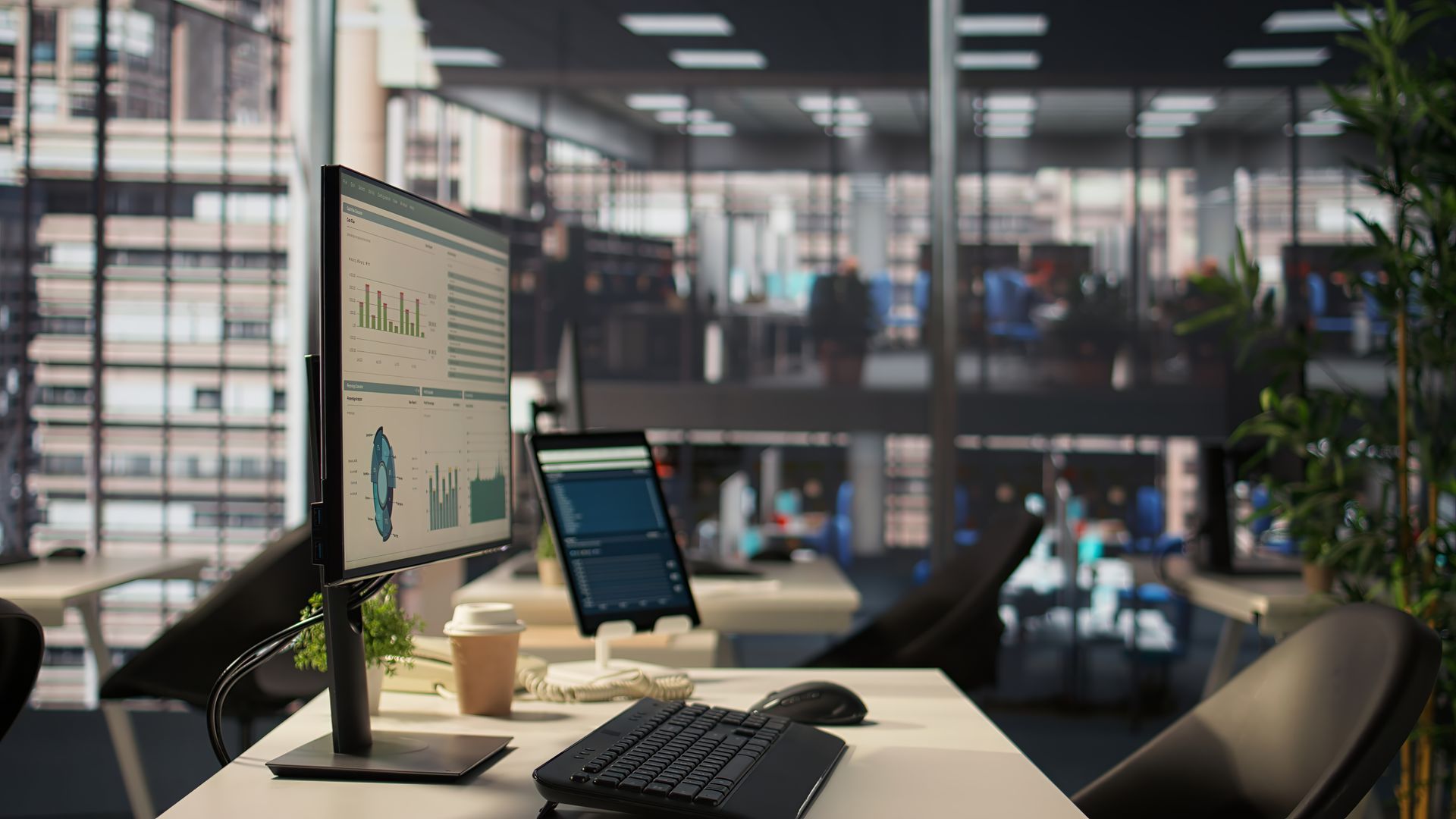 A desk with a computer monitor showing charts and a tablet in a modern, blurred office setting.