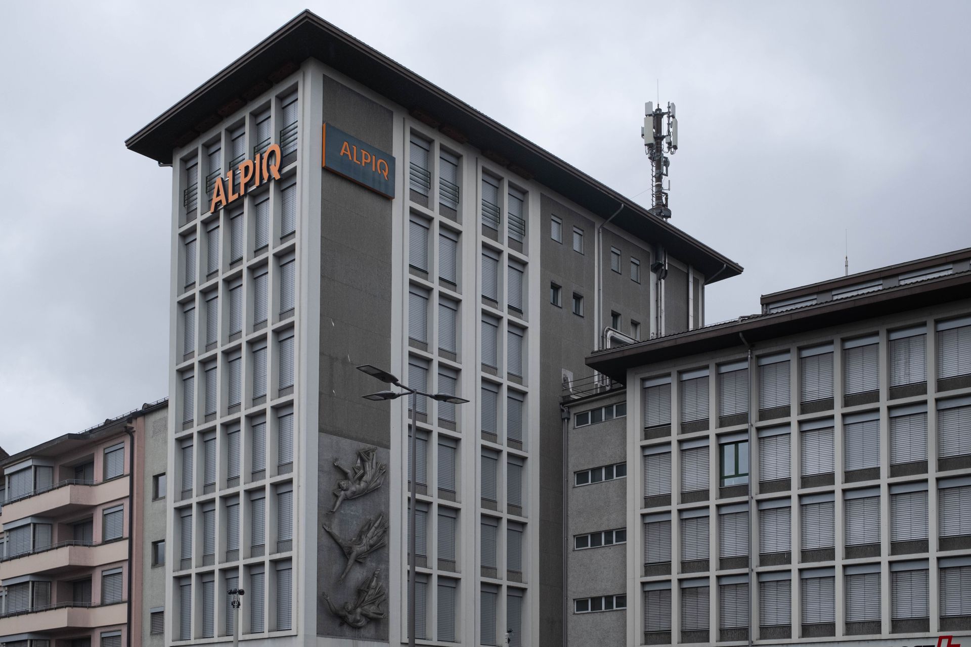 A multi-story gray administrative building with grid-pattern windows, an Alpin logo, and a rooftop cellular antenna.