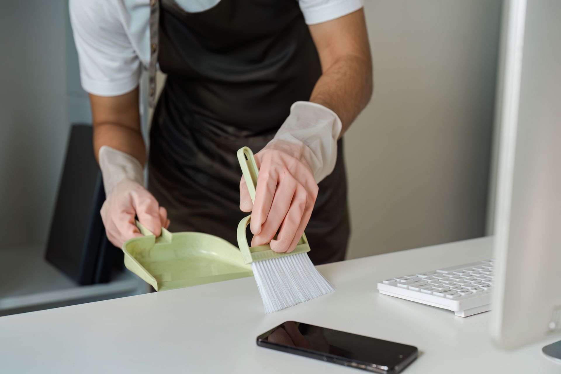 A person wearing gloves uses a small brush and dustpan to clean a white office desk next to a smartphone.