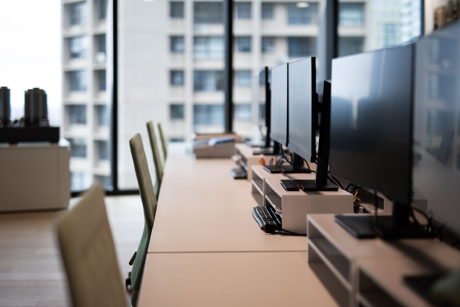 Row of desks with computer monitors in a modern office with large windows overlooking a city building.