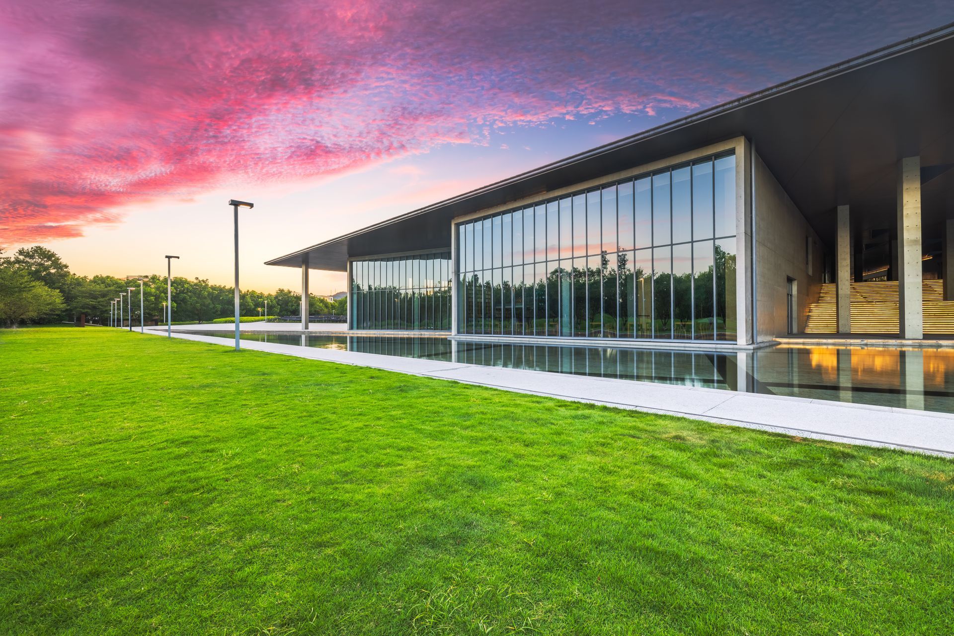 Modern building with glass walls beside a reflecting pool and grassy lawn under a vibrant sunset sky.