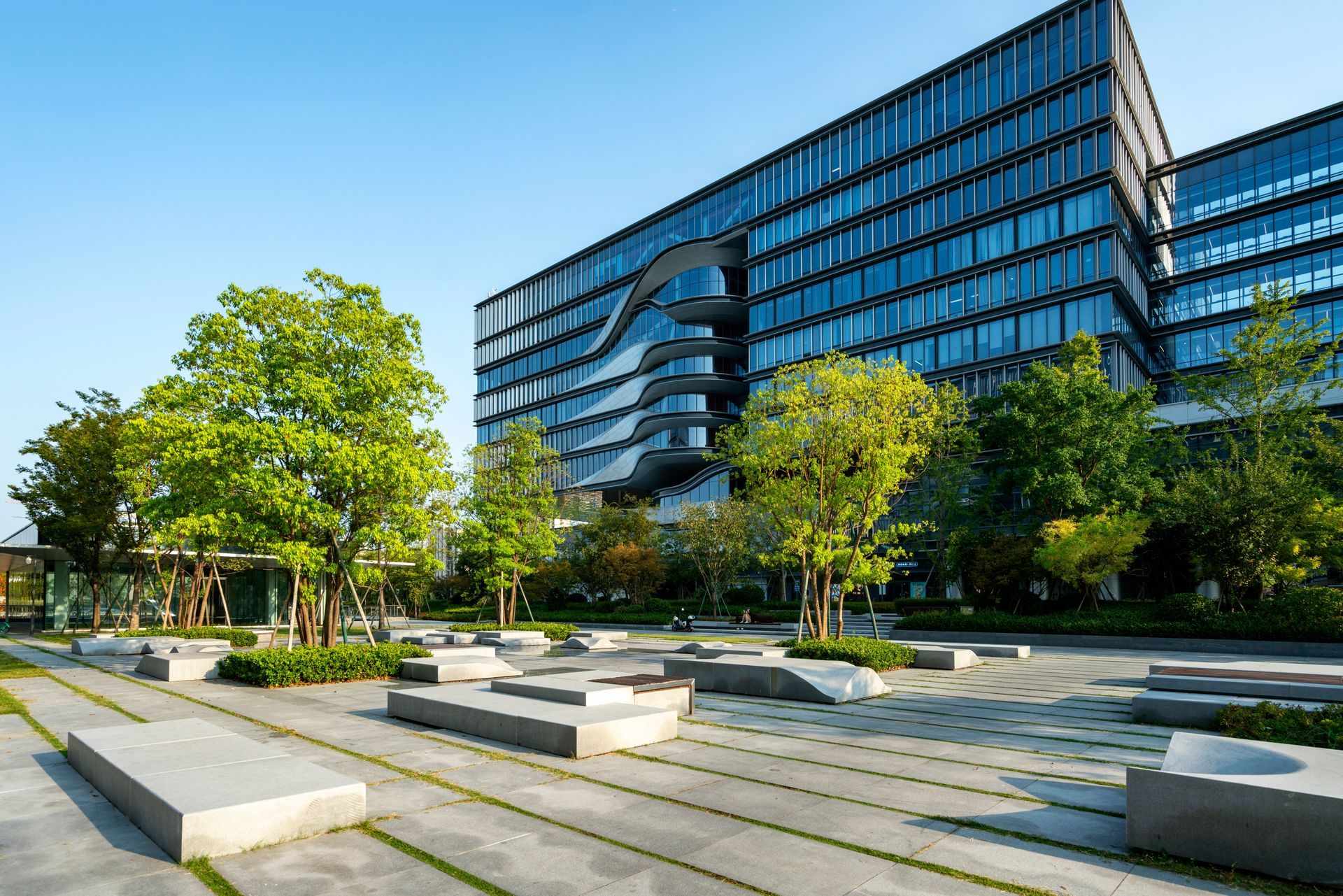 Modern office building with a curved glass facade overlooking a paved plaza with stone benches and green trees.