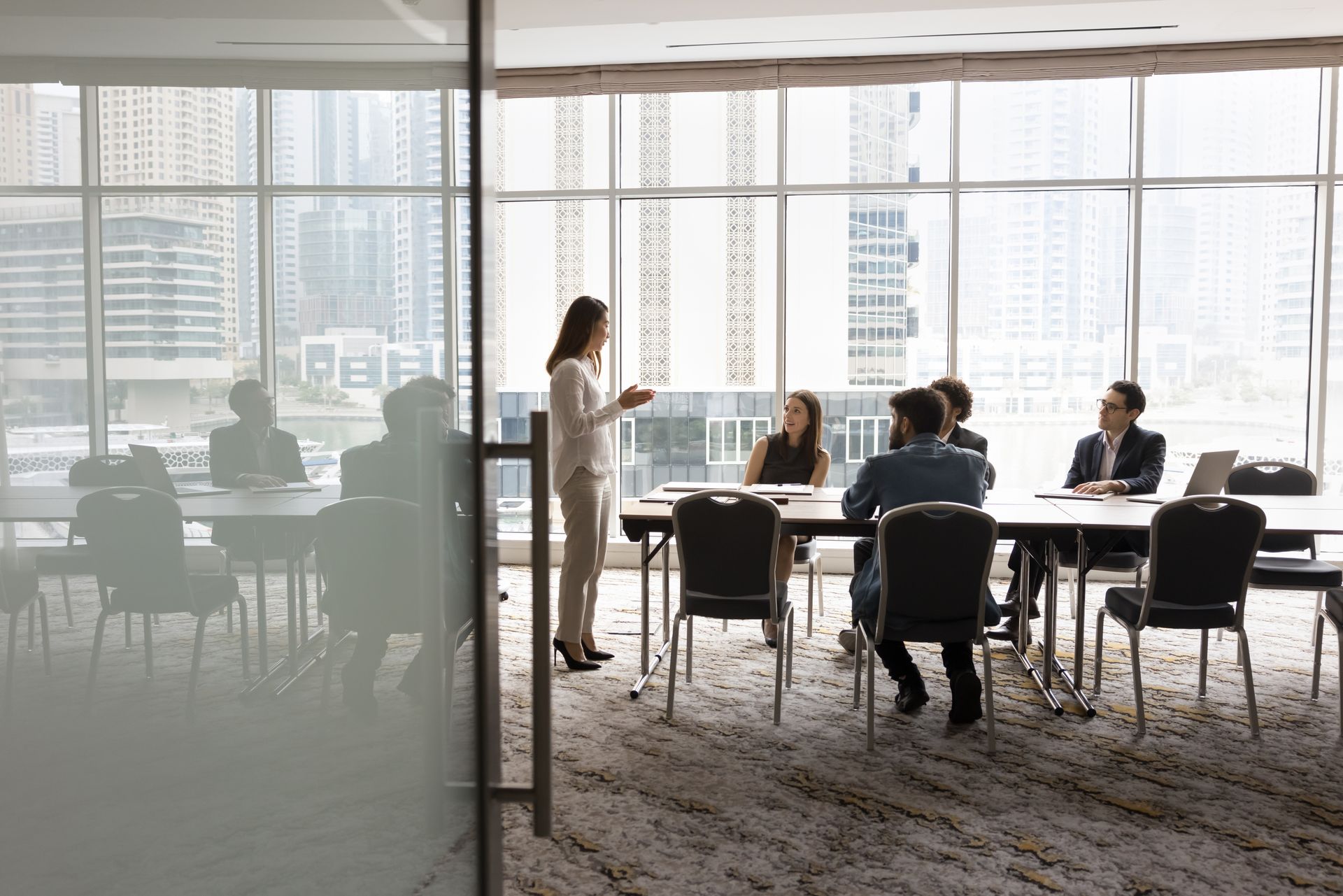A professional stands and leads a meeting with colleagues seated at a conference table in an office with city views.