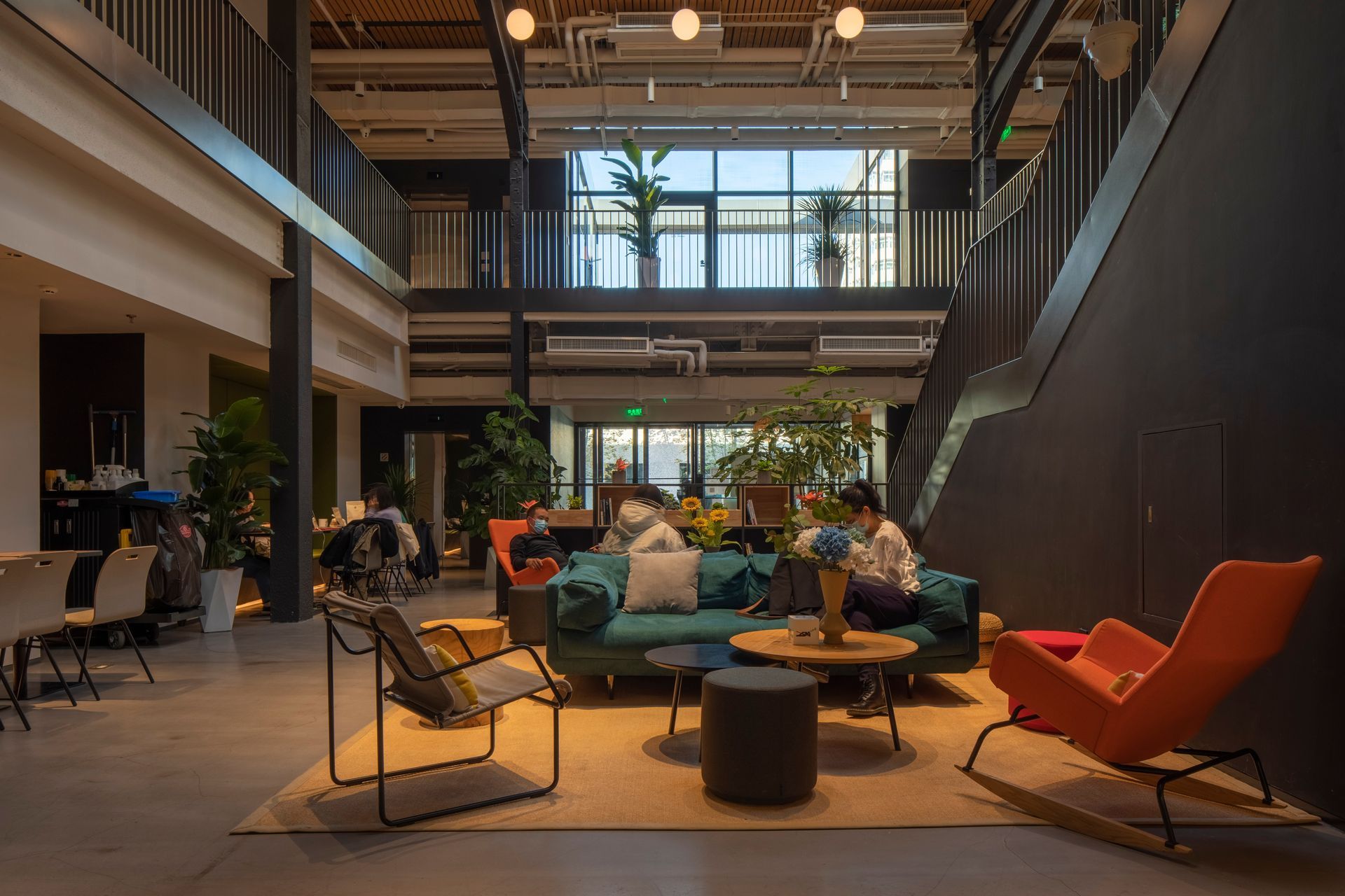 A modern, open-plan workspace featuring a green sofa, colorful chairs, and desks in a two-story industrial interior.