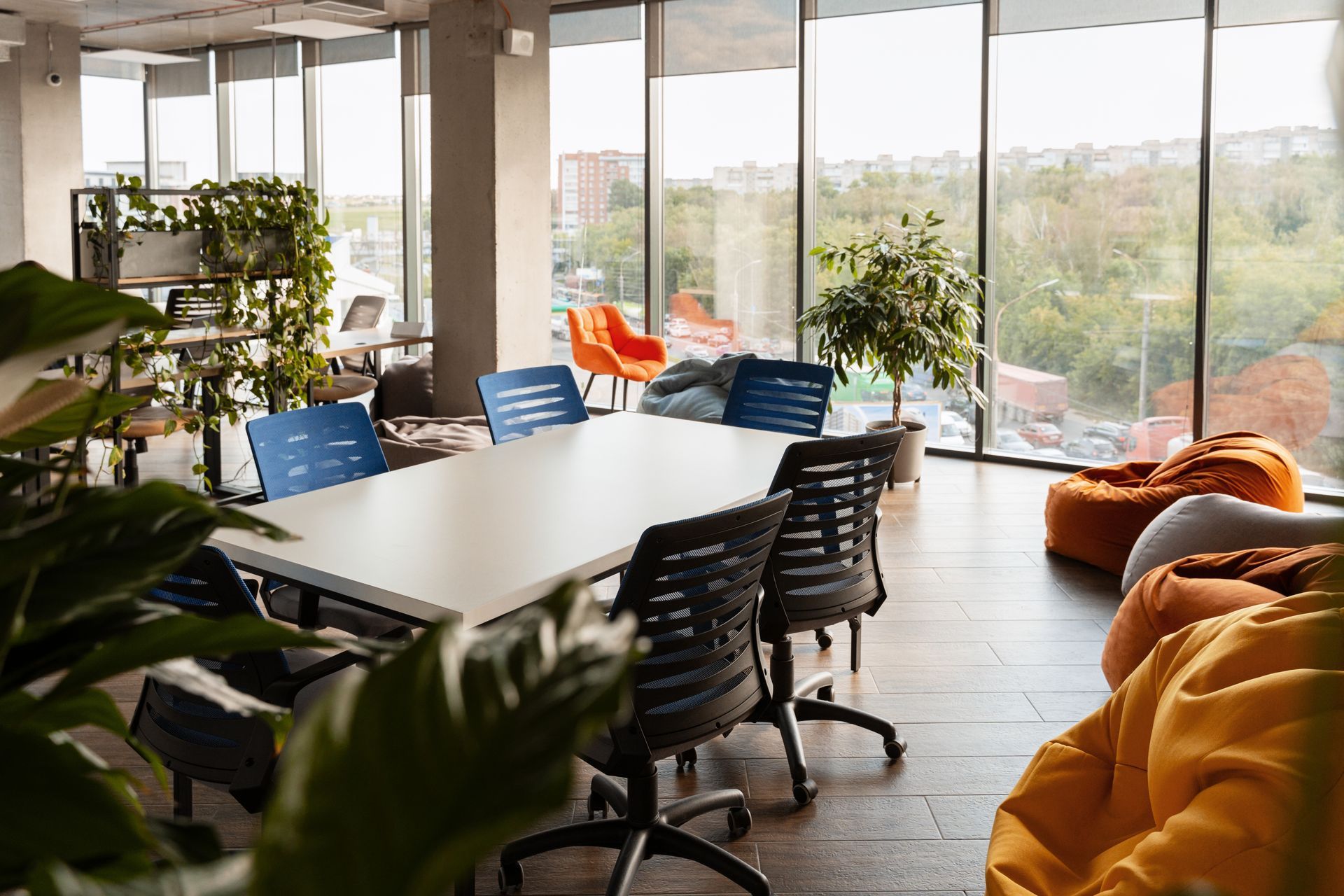 Modern office meeting room with a large white table, dark ergonomic chairs, floor-to-ceiling windows, and bean bag chairs.