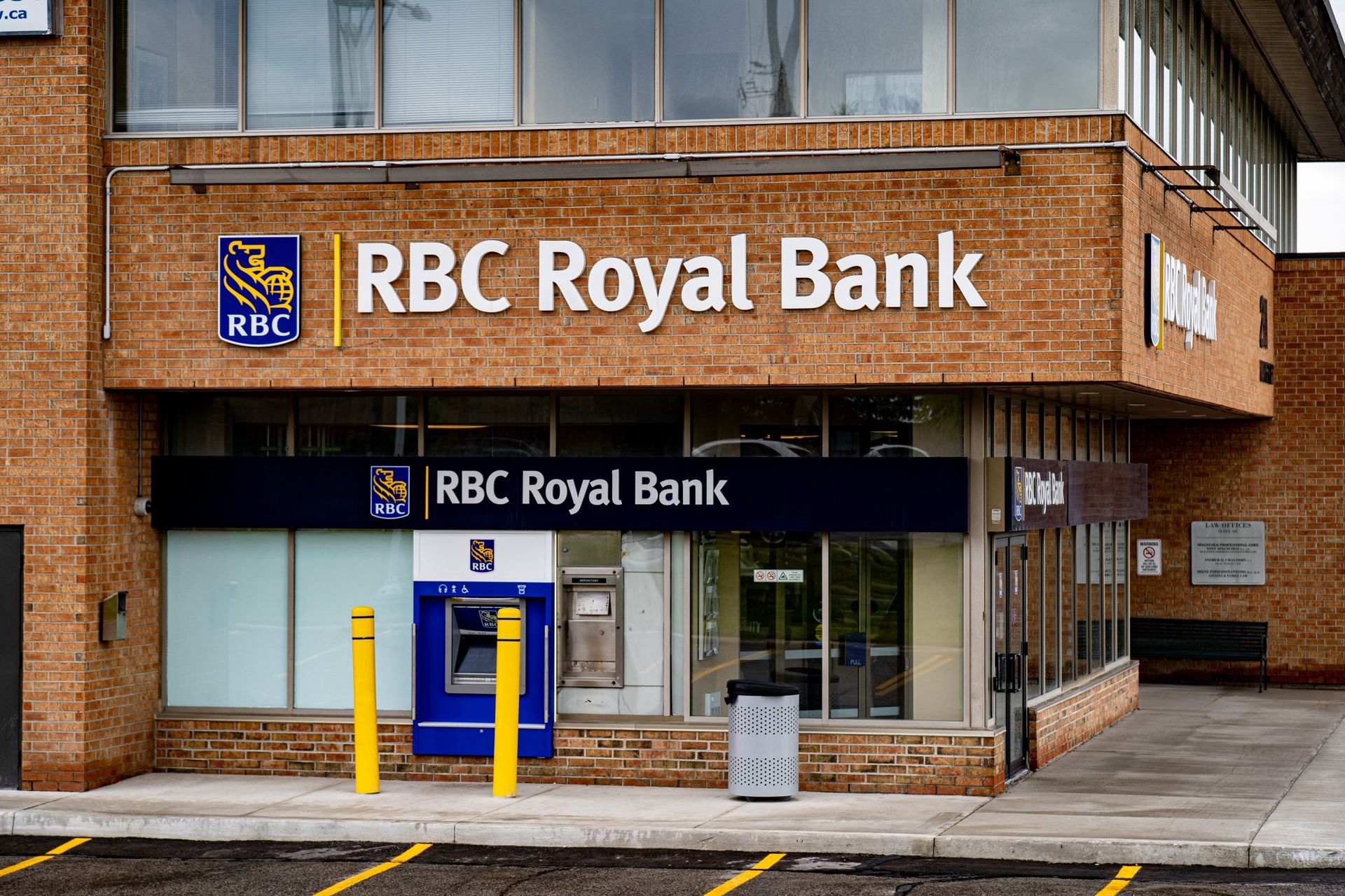 A brick RBC Royal Bank branch with a walk-up ATM, glass storefront, and yellow bollards in the parking lot.