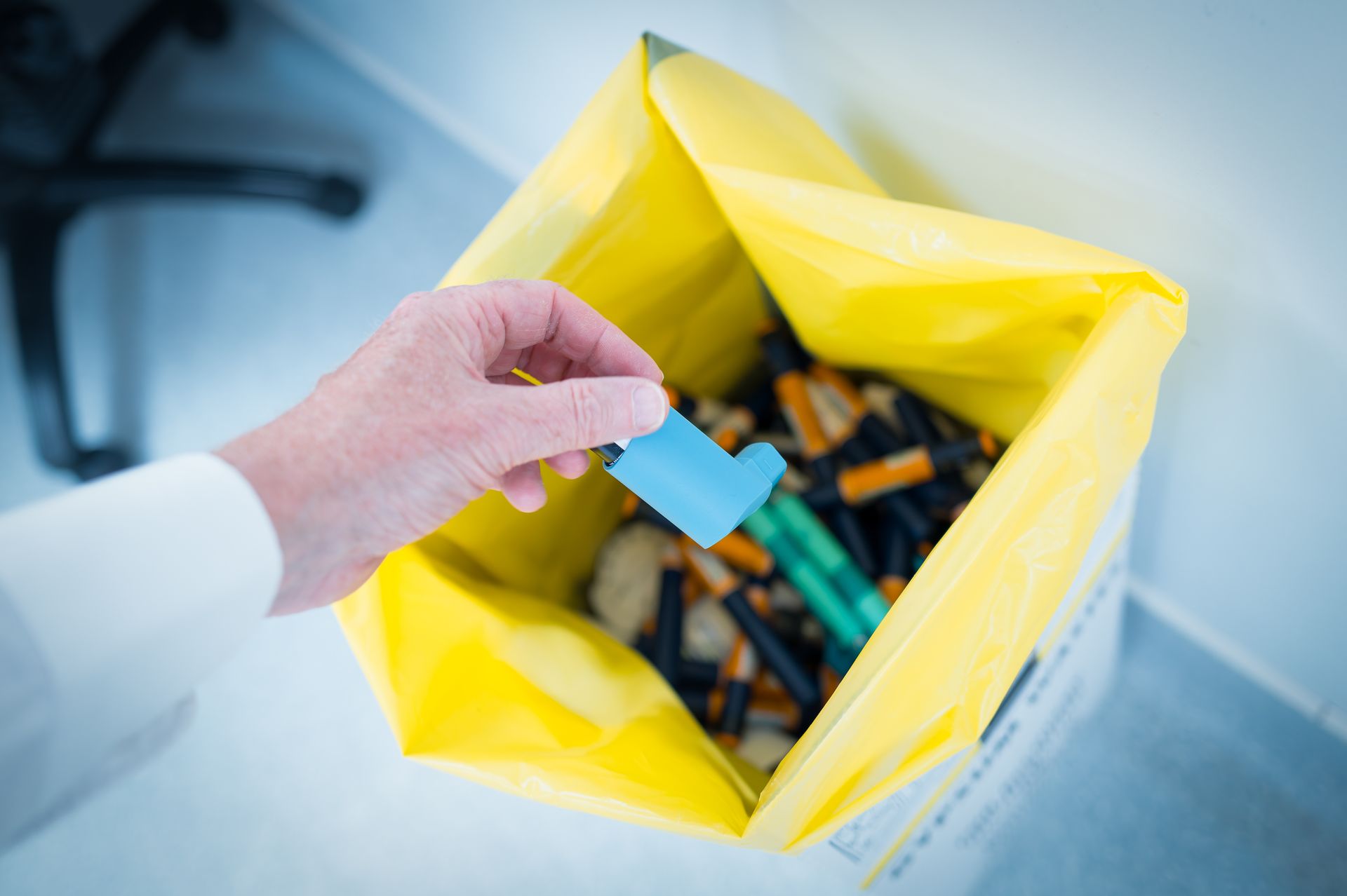 A hand holds a blue inhaler over a yellow clinical waste bag filled with used medical devices.