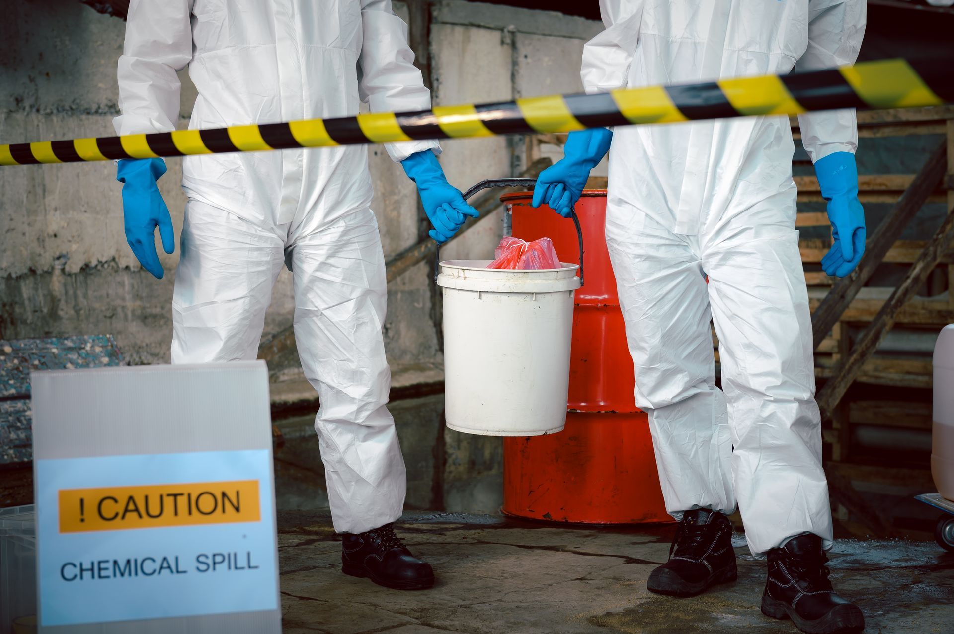 Two workers in white hazmat suits and blue gloves stand behind caution tape near a white bucket and orange drum.