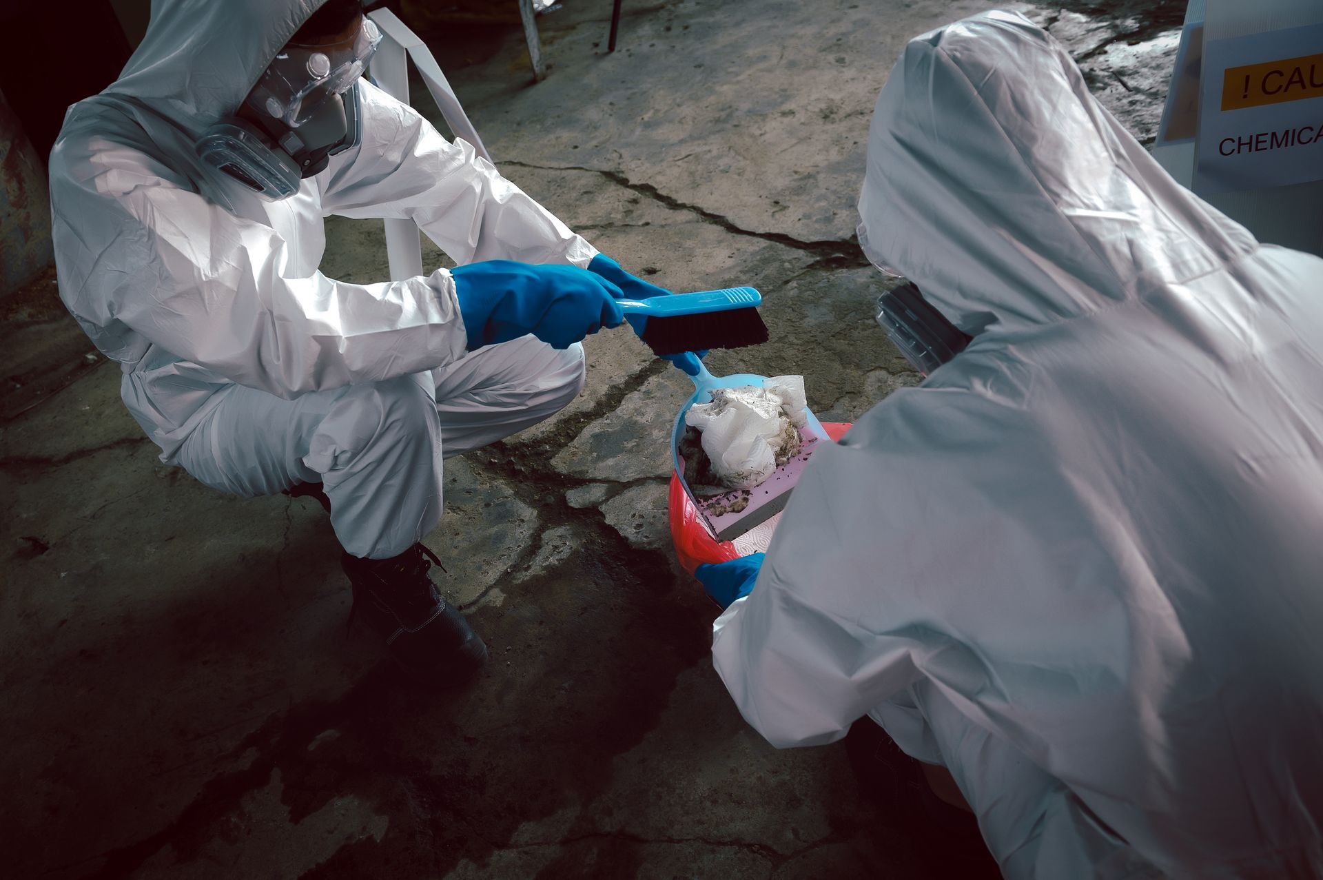 Two people in white protective suits and masks crouch on a cracked surface, using brushes to collect debris into a bucket.