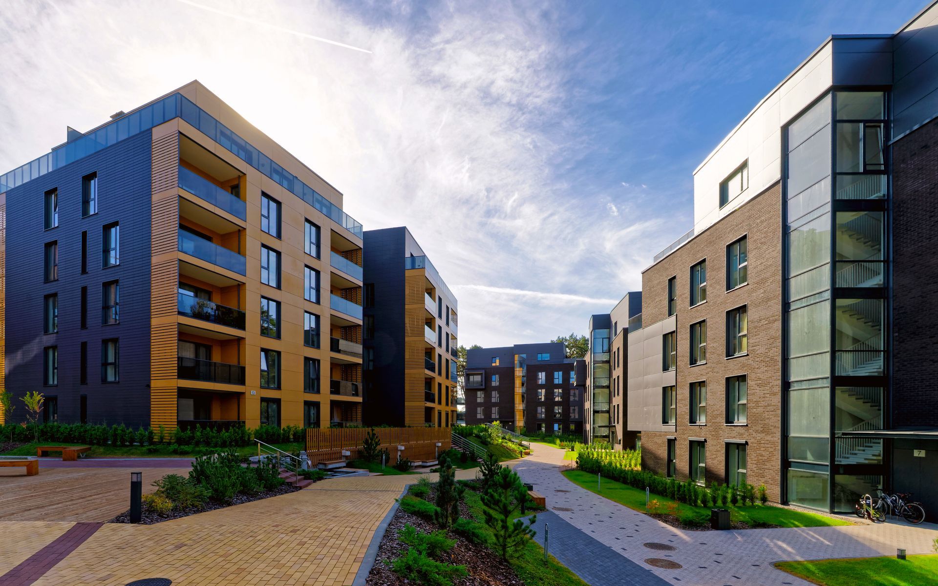 Modern apartment buildings with dark brick and wood accents surround a paved courtyard with greenery under a bright sky.
