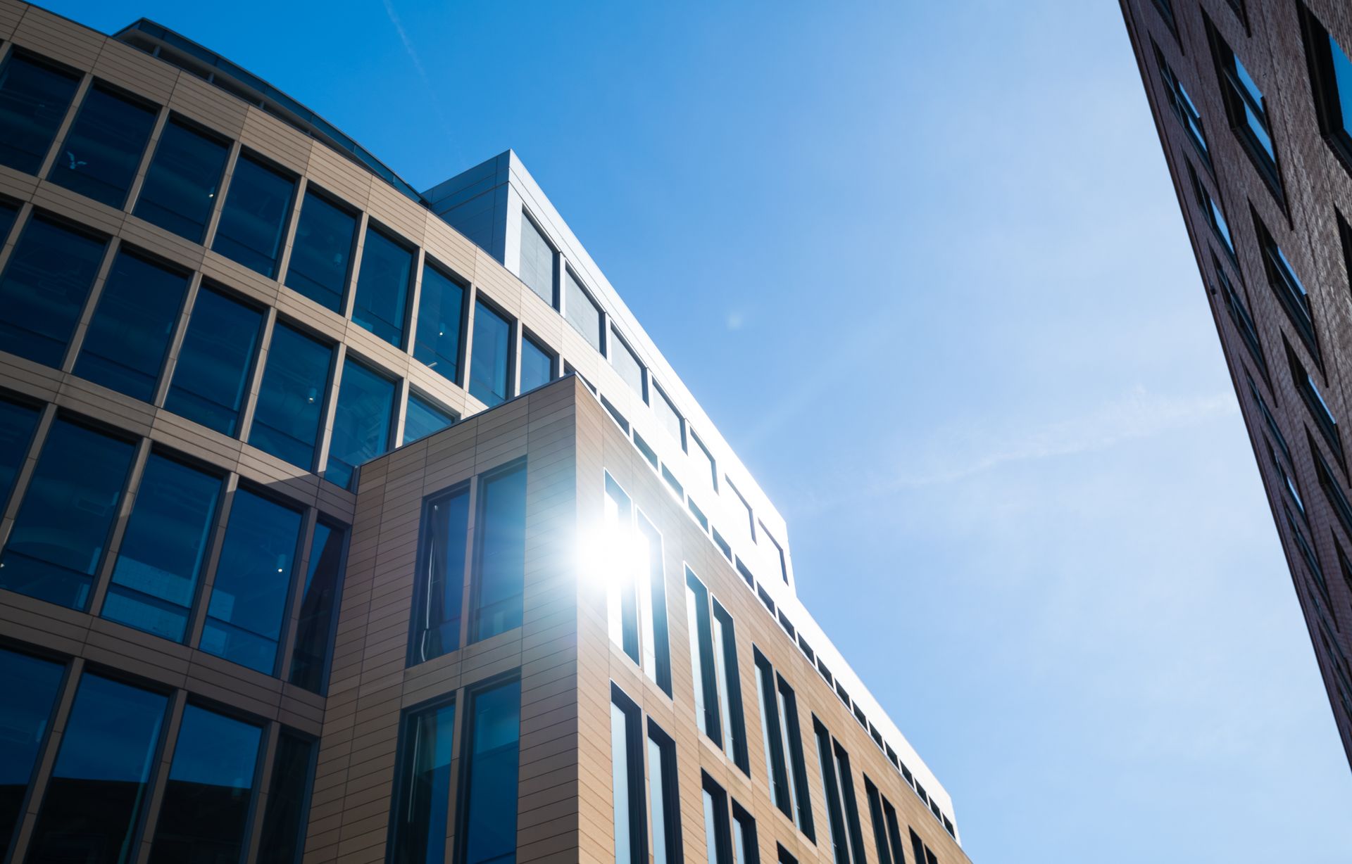 A low-angle view of modern, tan-brick office buildings against a bright, clear blue sky with a sun flare.