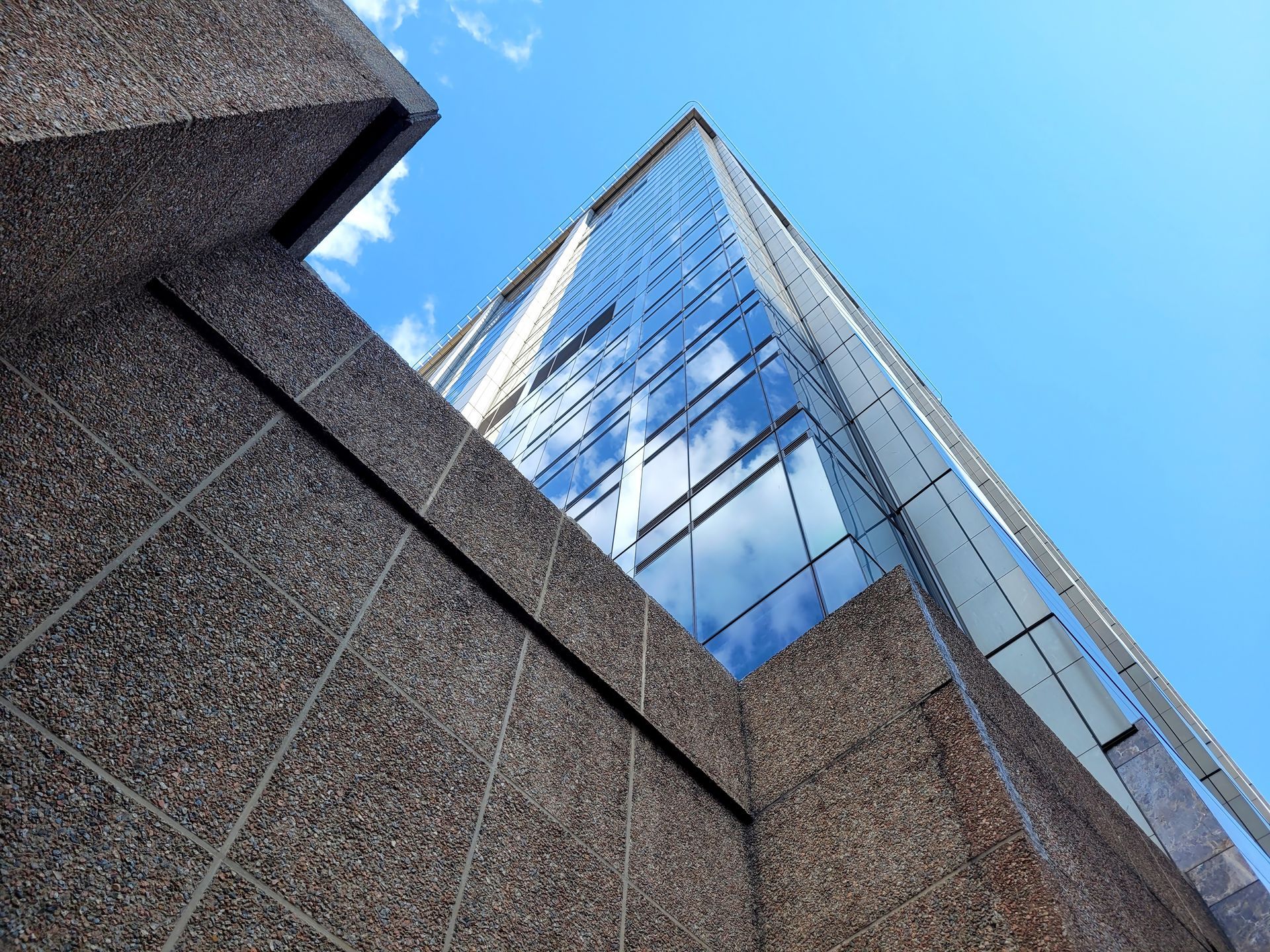 A low-angle view of a modern glass skyscraper reflecting the blue sky, juxtaposed with a textured concrete wall.