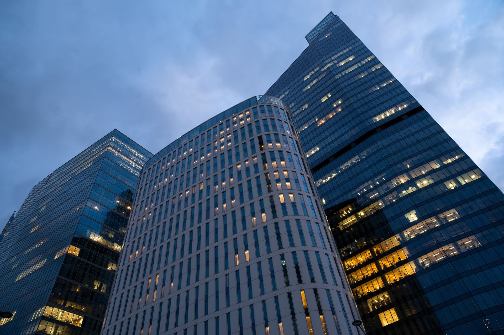Low-angle view of three modern skyscrapers against a cloudy blue twilight sky, with lights visible in many windows.