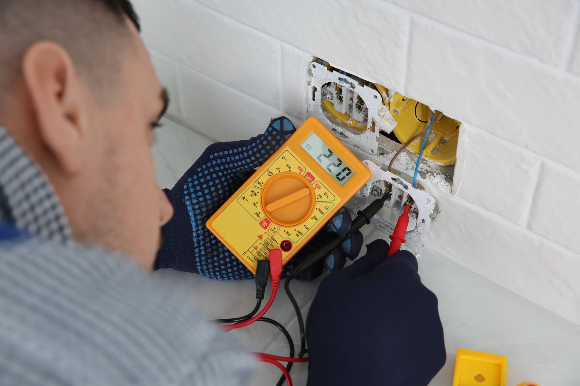 An electrician wearing gloves uses a yellow multimeter to test the voltage of an open electrical wall outlet.