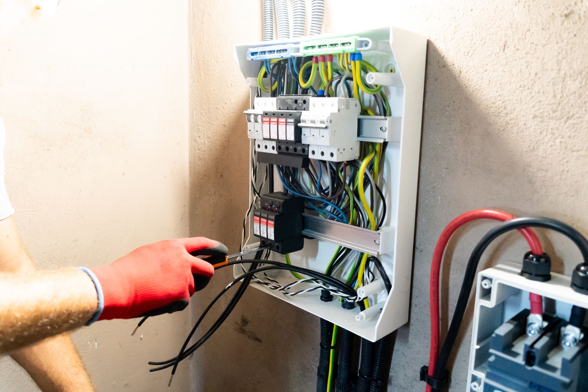 An electrician in red gloves wires a white electrical panel on a wall near a second junction box.