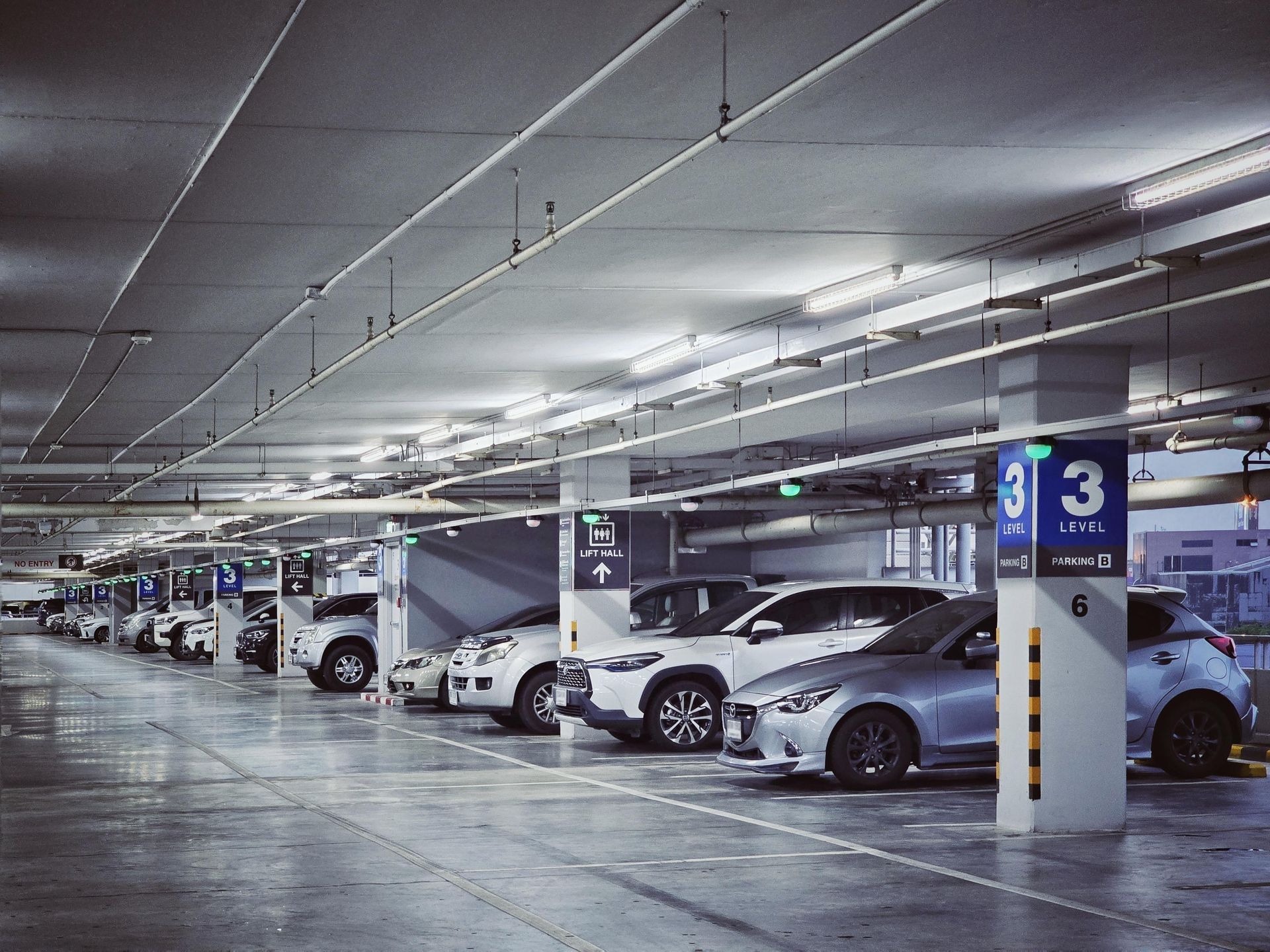 A line of cars parked in a brightly lit indoor parking garage with concrete floors and labeled structural pillars.