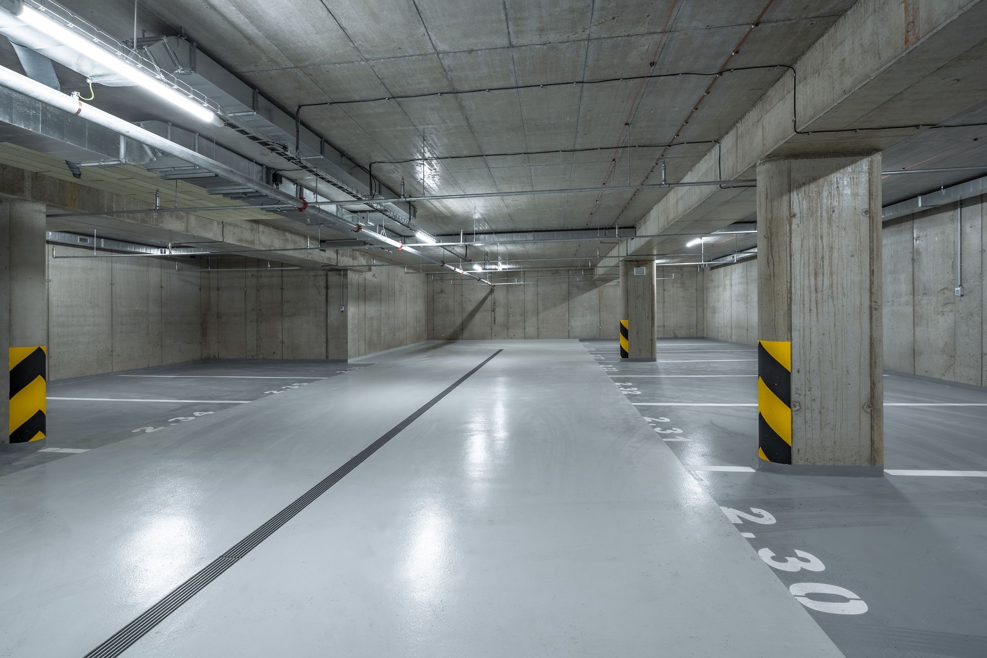 An empty, modern underground parking garage with gray concrete walls, pillars with yellow and black tape, and marked spots.