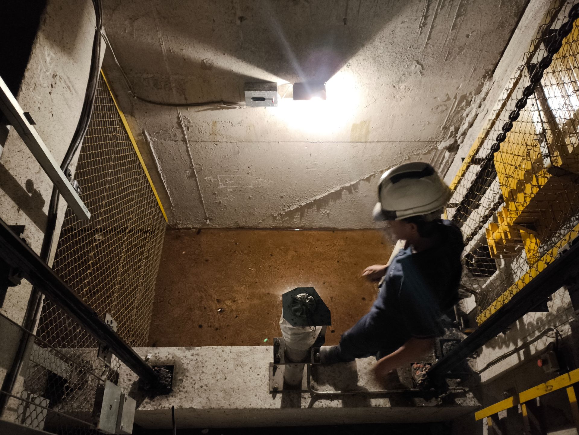 A worker in a hard hat inspects an open elevator shaft from an elevated concrete platform.