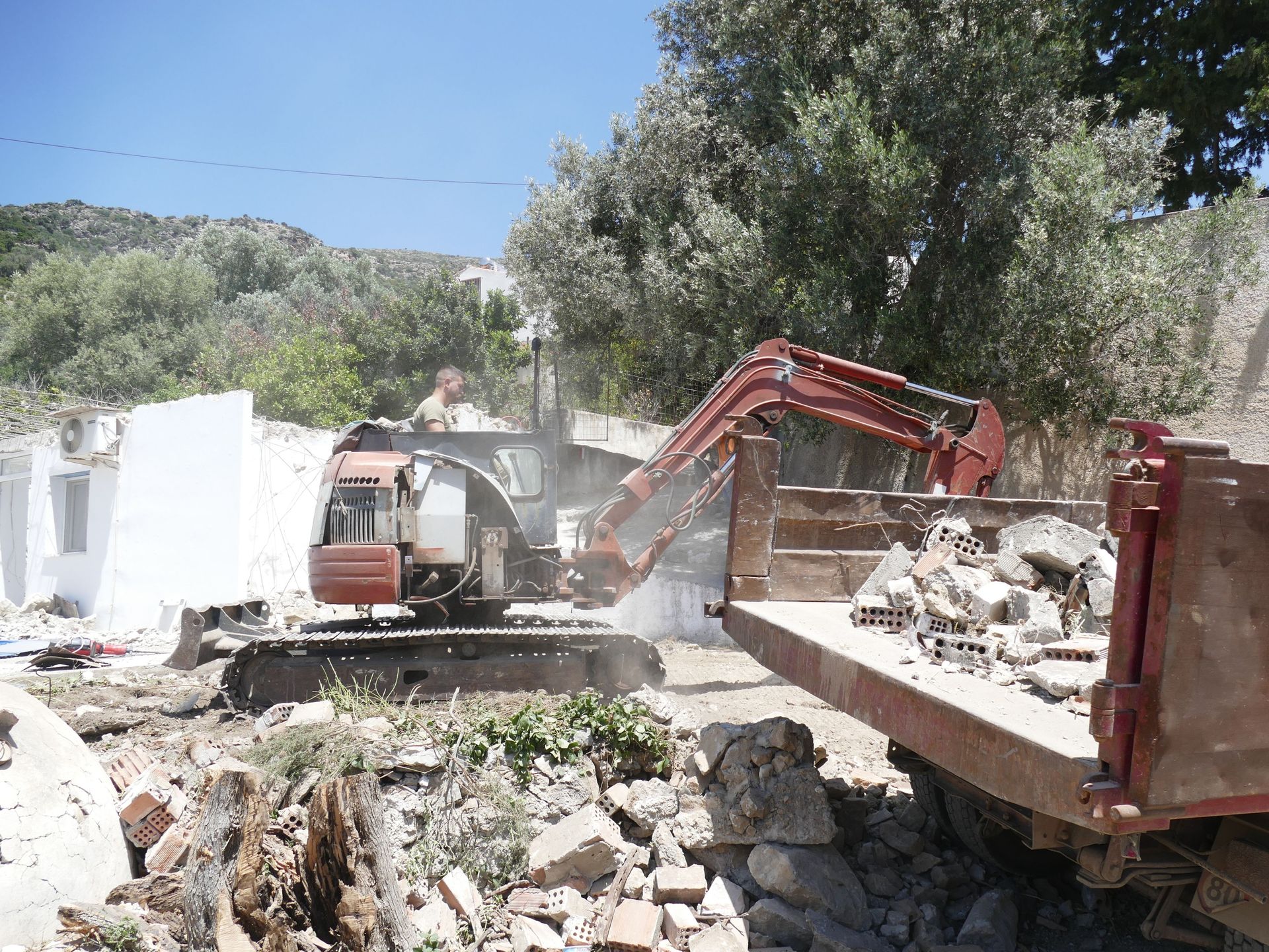 A red excavator loads rubble into a dump truck at a demolition site near trees and white buildings under a blue sky.