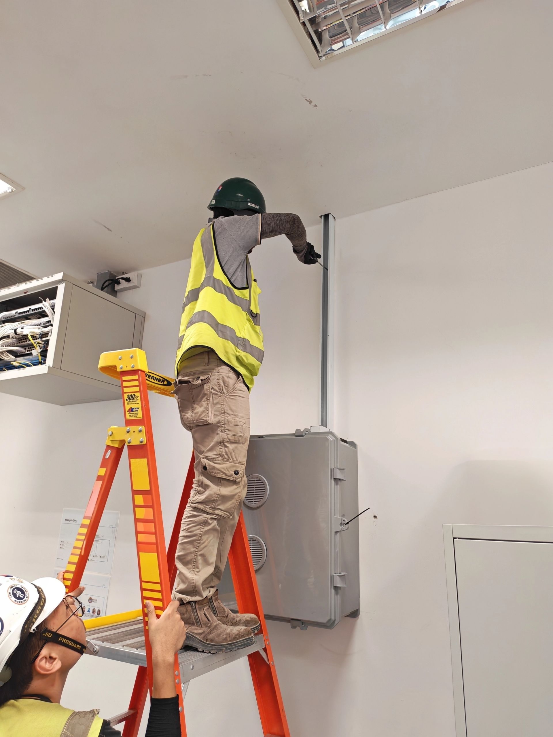 Worker on ladder installing a ceiling fixture beside an electrical panel in an indoor room