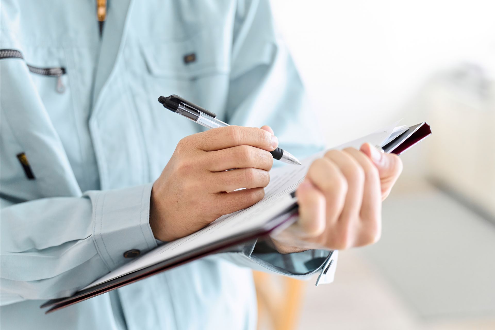 A person in a light blue work shirt writes on a clipboard held in their hand.