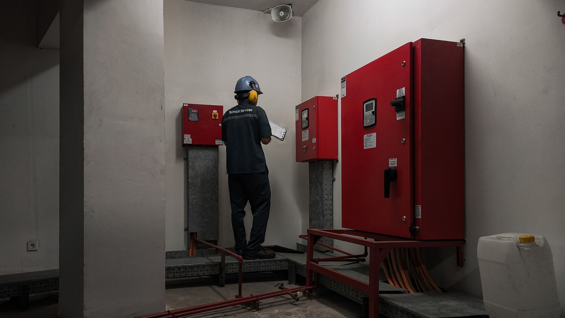 A technician in a hard hat and ear protection inspects red industrial electrical cabinets in a plain, white room.