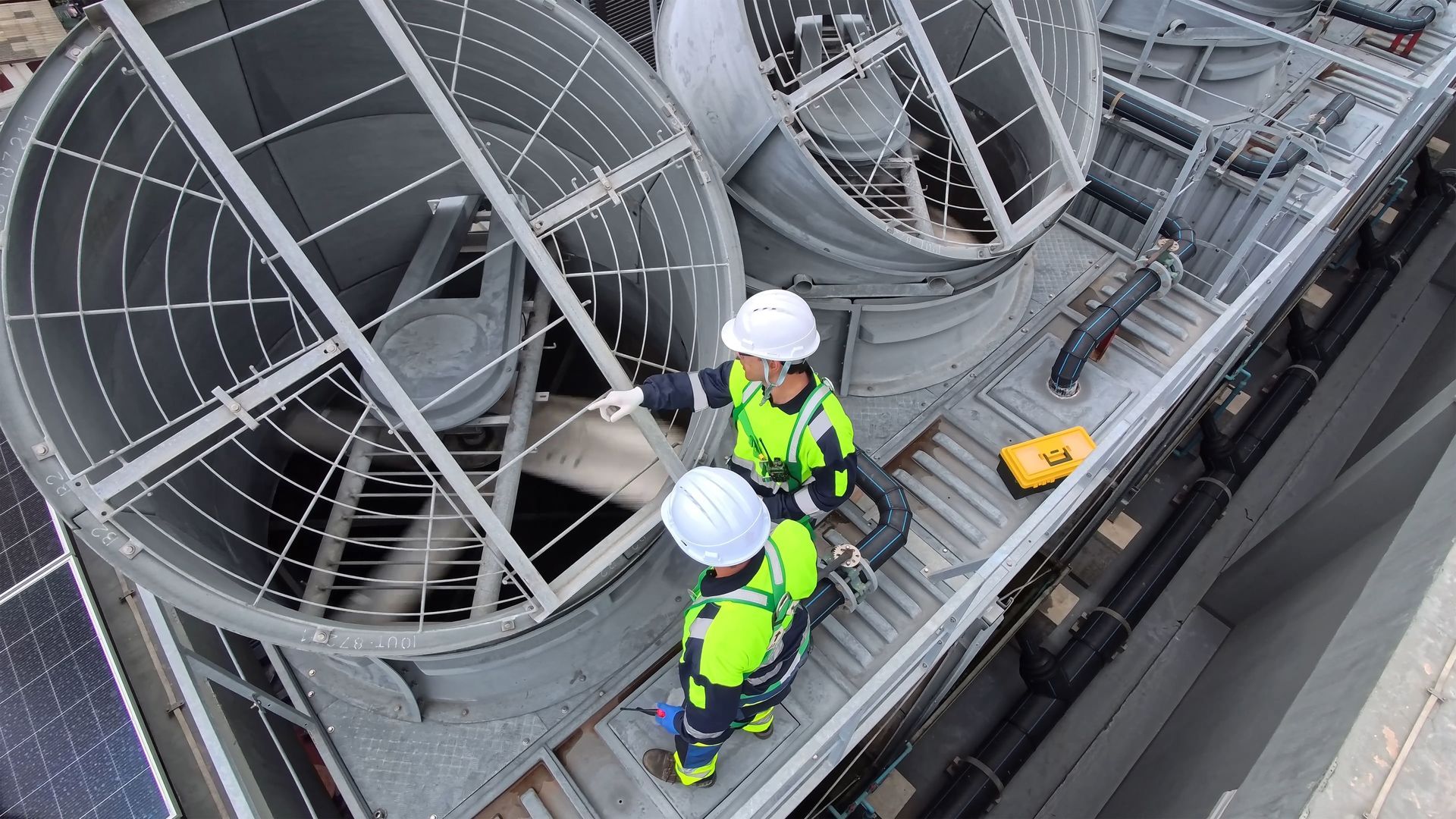 Two workers in high-visibility safety gear and hard hats inspect a large industrial cooling fan on a rooftop.