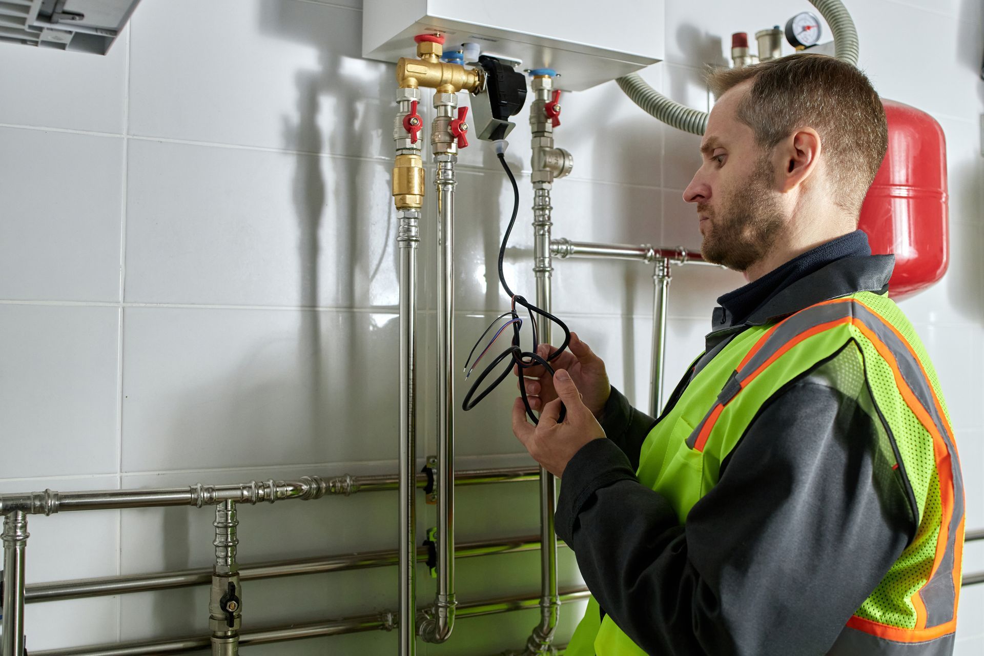 A person in a high-visibility vest inspects electrical wiring connected to pipes beneath a wall-mounted boiler.