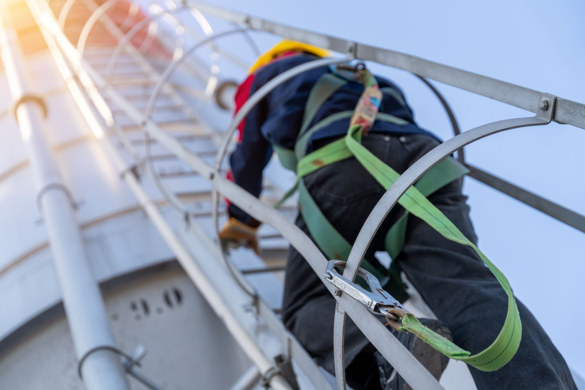 A worker in a safety harness and hard hat climbs a vertical industrial ladder on a large cylindrical structure.