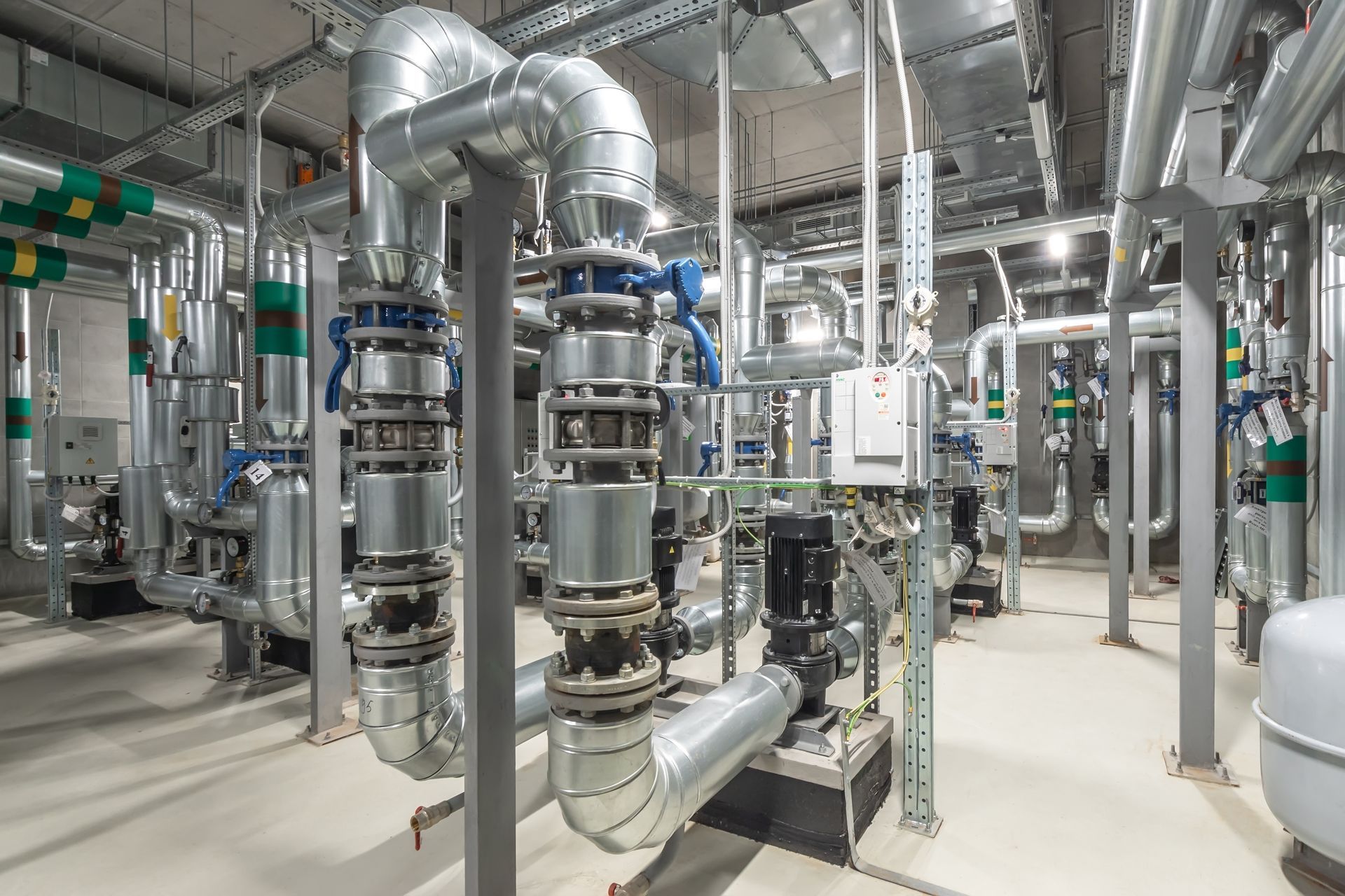 A mechanical room featuring complex insulated metal pipes, valves, and industrial pump systems on a concrete floor.