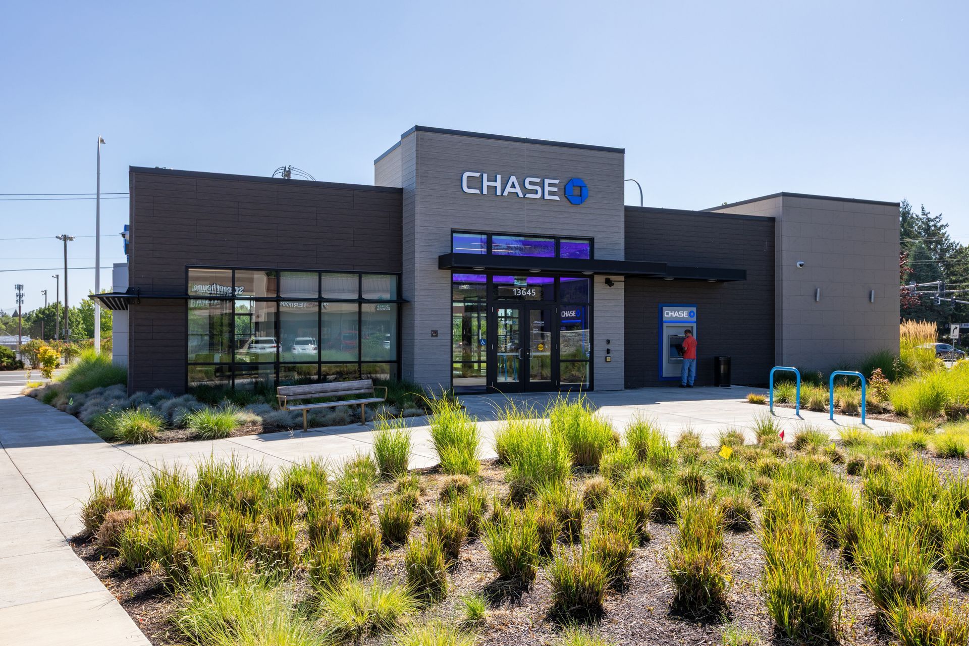 A modern Chase bank branch with a dark gray exterior, glass entrance, and surrounding landscaping under a blue sky.