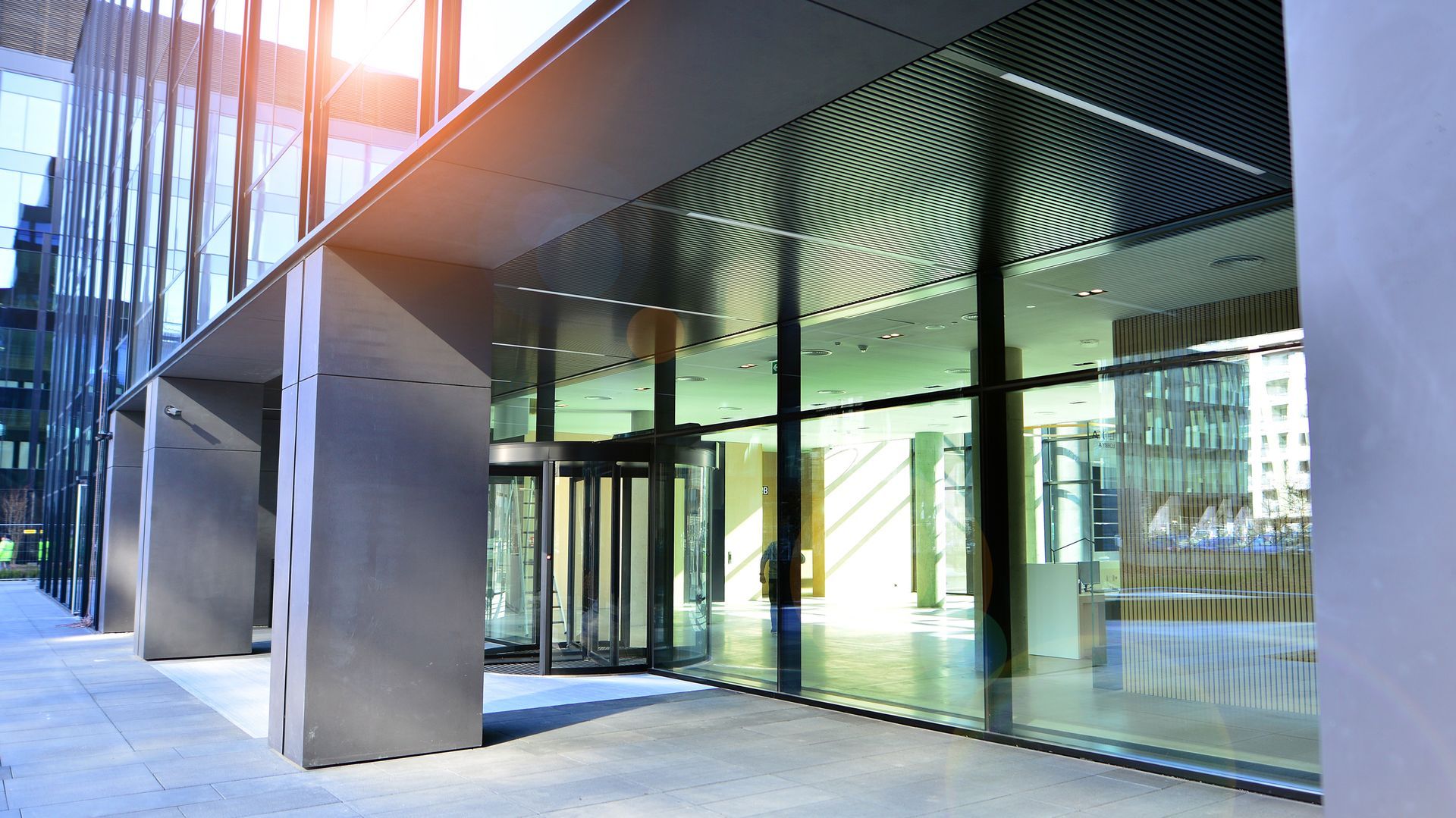 Modern building entrance with a glass facade, revolving doors, and a covered walkway under bright sunlight.