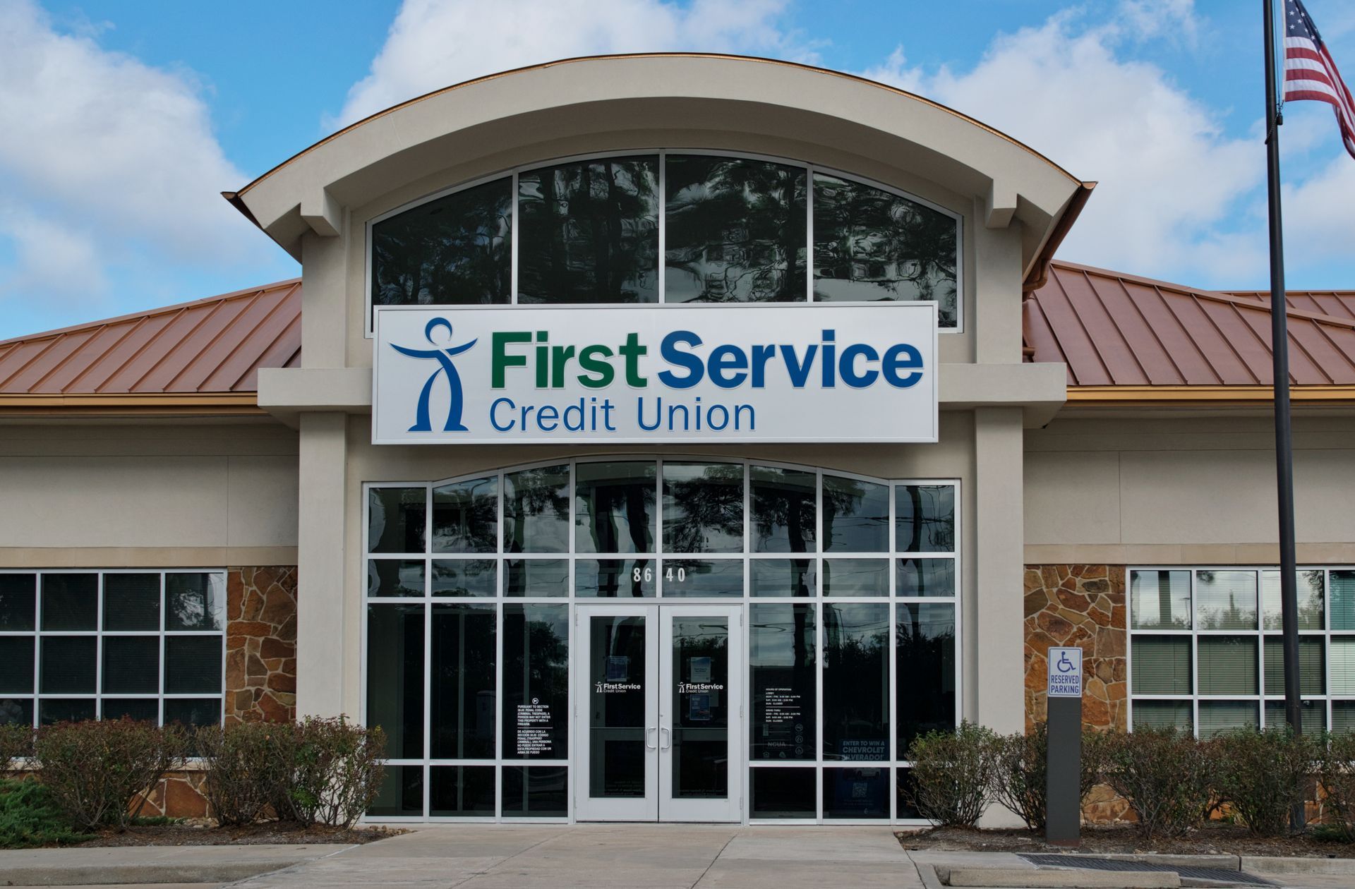 The exterior of a First Service Credit Union branch featuring a white sign with a logo, glass doors, and an American flag.