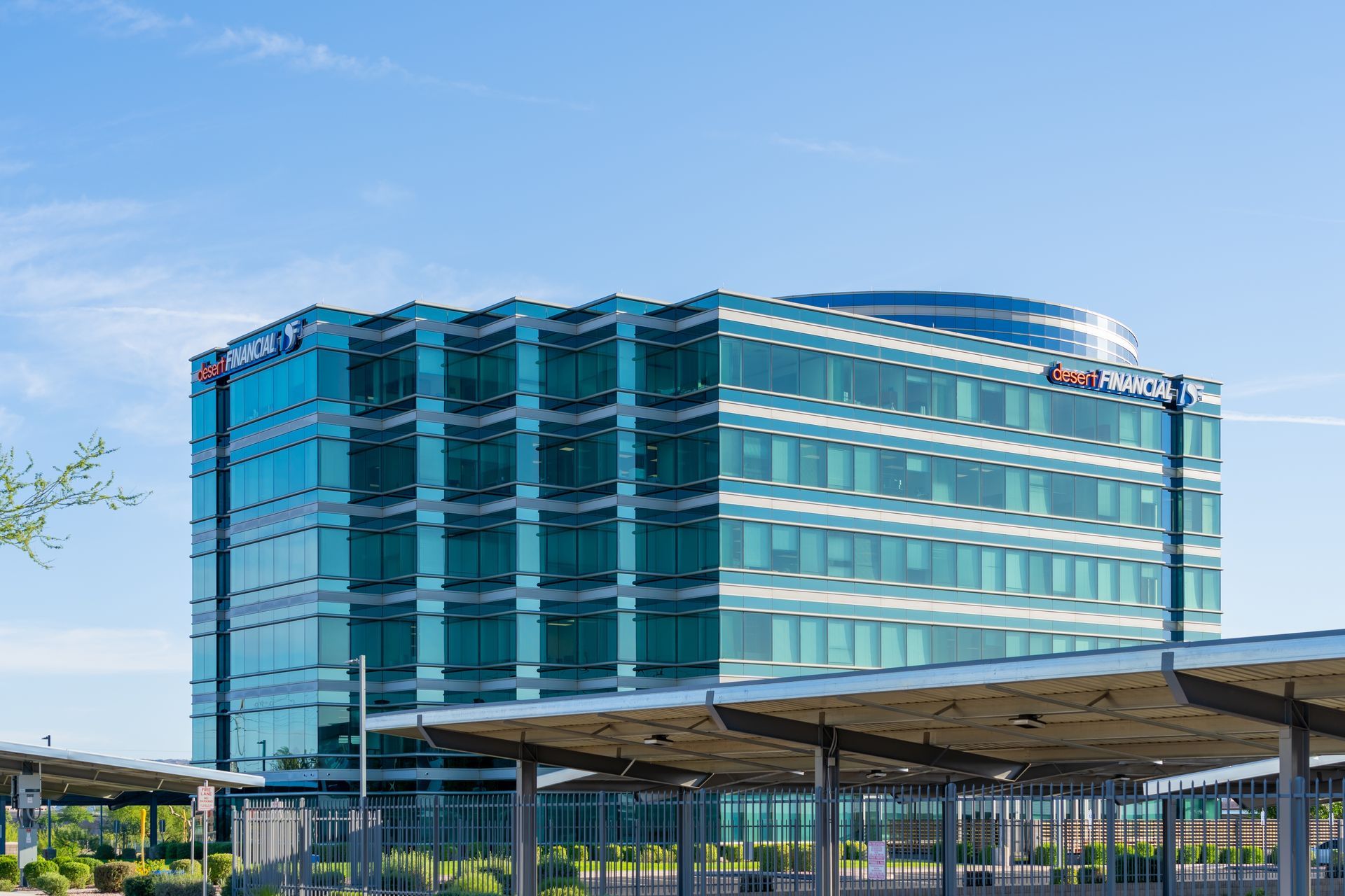 A modern, multi-story blue-glass office building under a clear sky, viewed from a parking area with shaded structures.