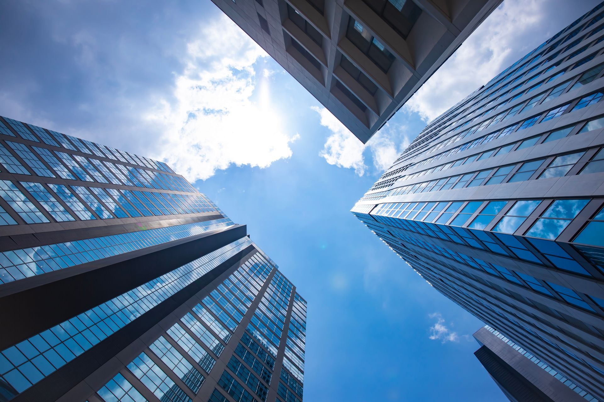A low-angle view of several modern glass skyscrapers stretching toward a blue sky with soft white clouds.