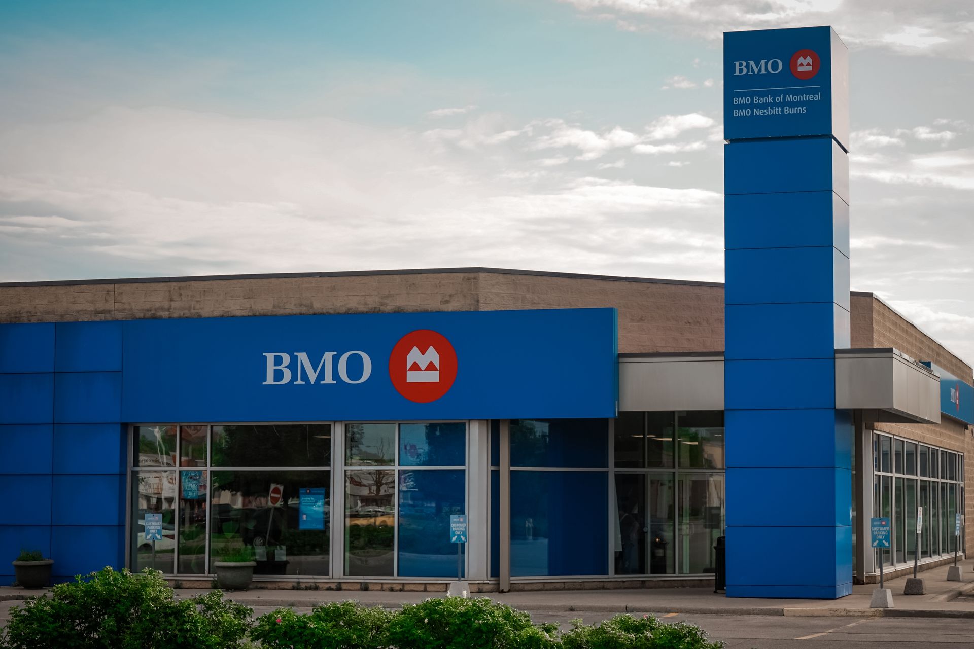 A BMO bank branch building with blue exterior signage and a tall, rectangular blue pillar sign against a cloudy sky.