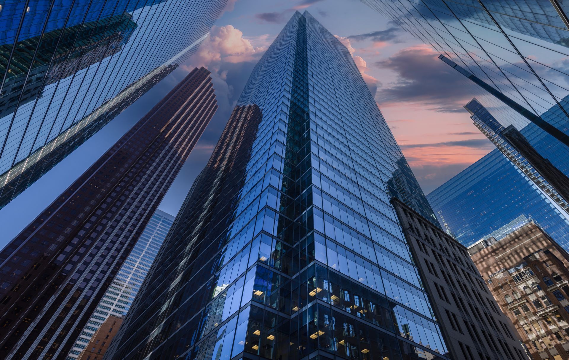 A low-angle view of multiple modern glass skyscrapers reaching toward a dusk sky with soft orange and blue tones.