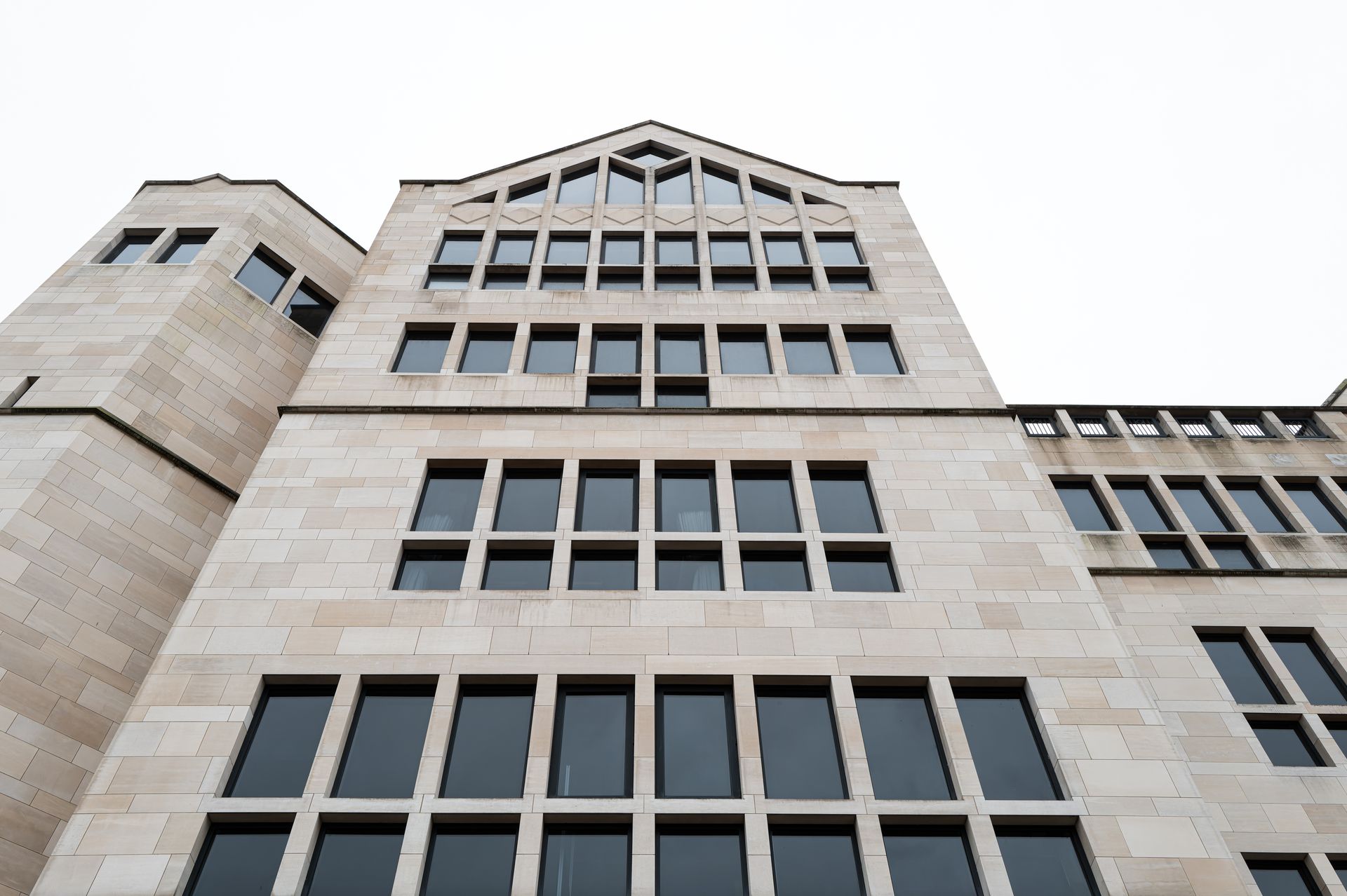 Low-angle view of a modern building facade featuring light-colored stone, rectangular grid windows, and a peaked roof.